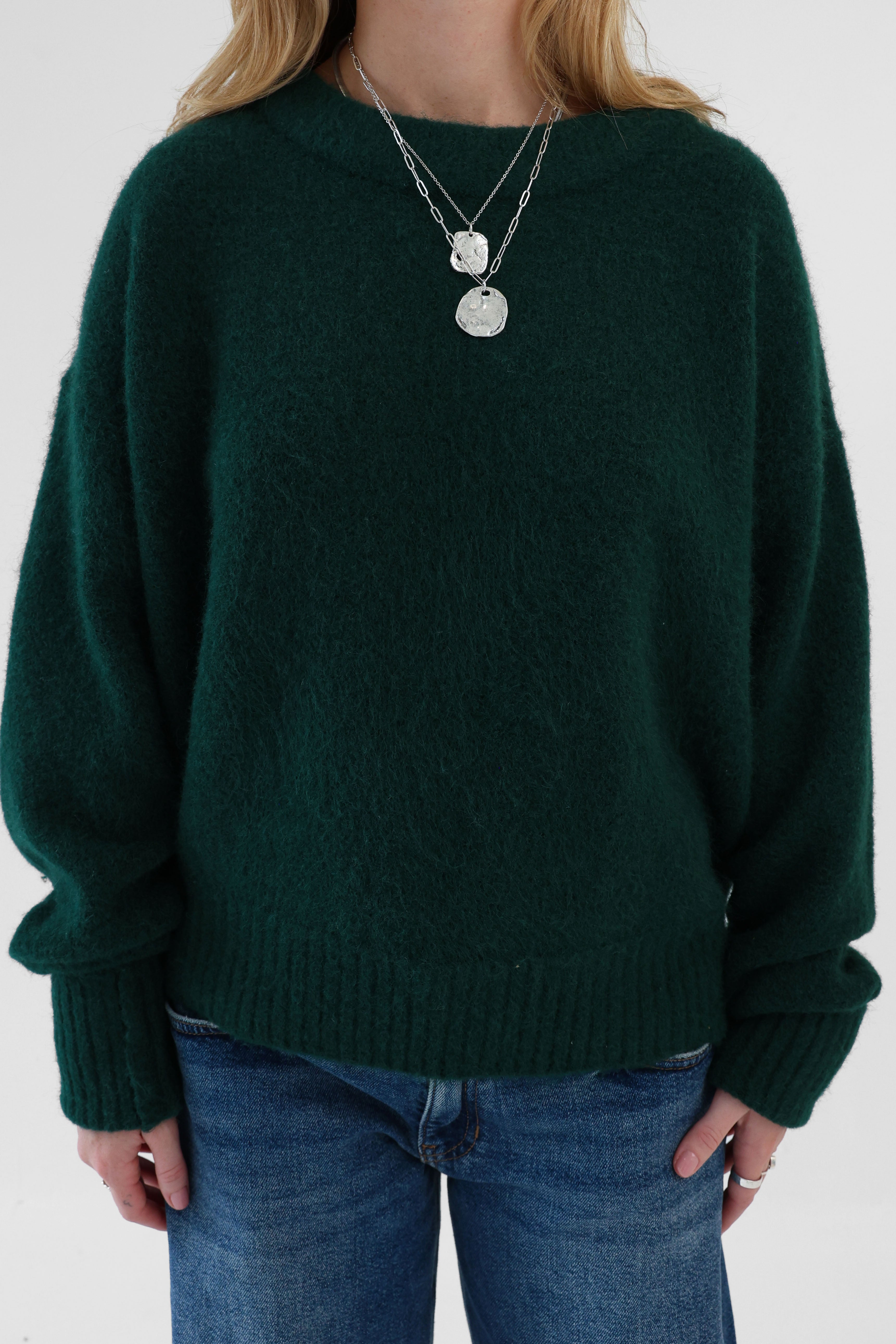 Person wearing a dark green sweater and blue jeans with a plain background
