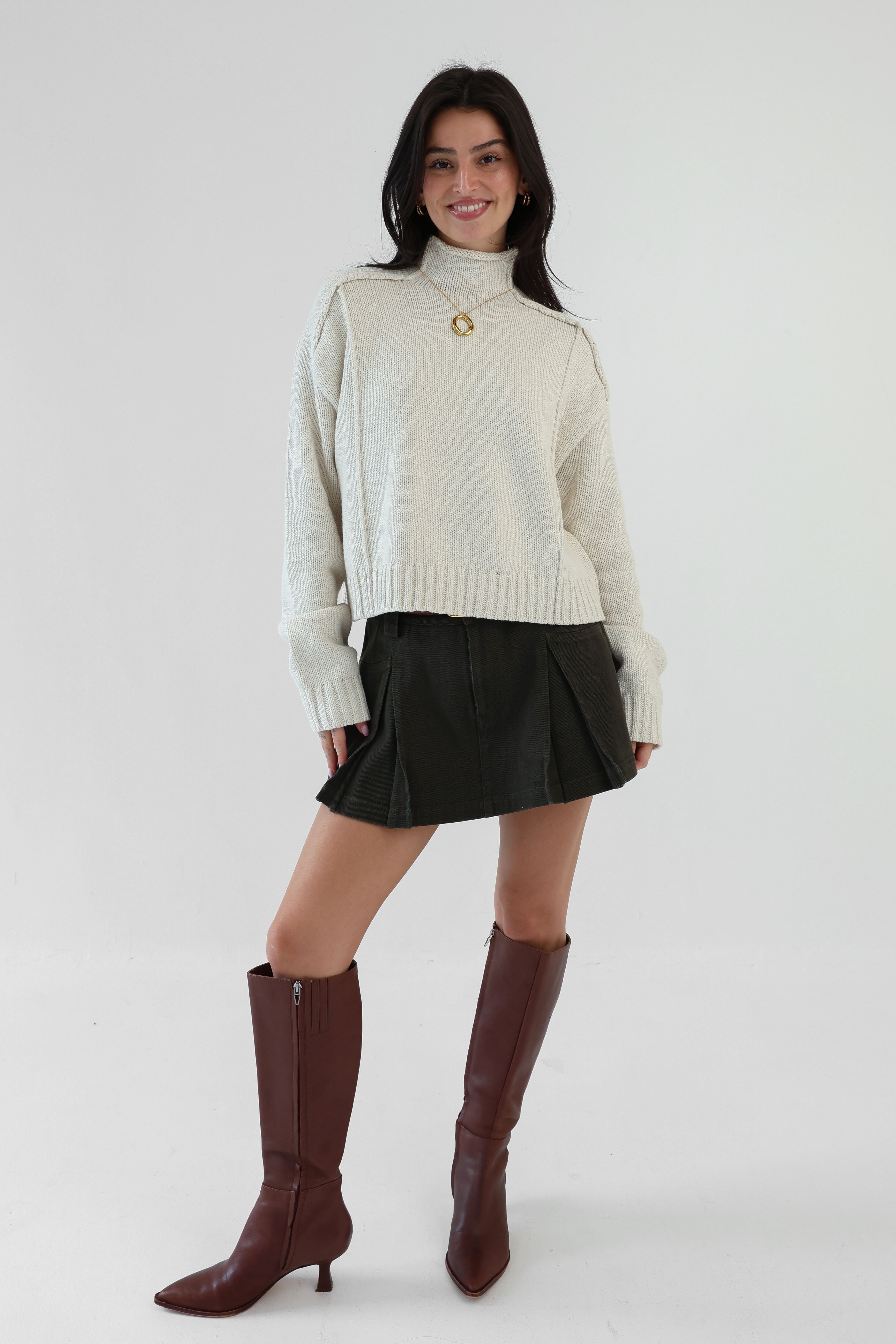 Woman wearing a cream sweater, black skirt, and brown knee-high boots on a white background