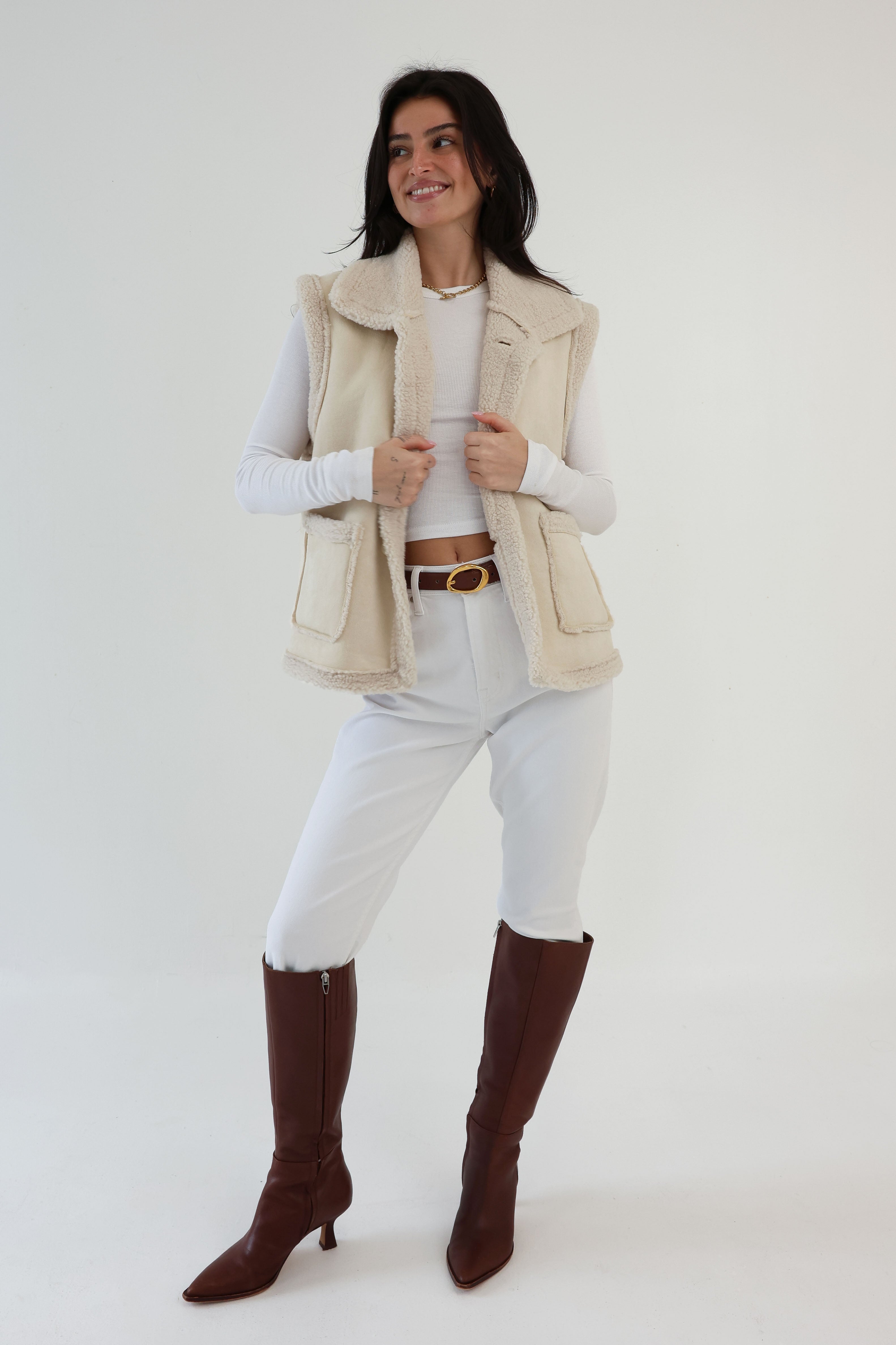 Woman wearing a beige vest, white pants, and brown boots on a white background