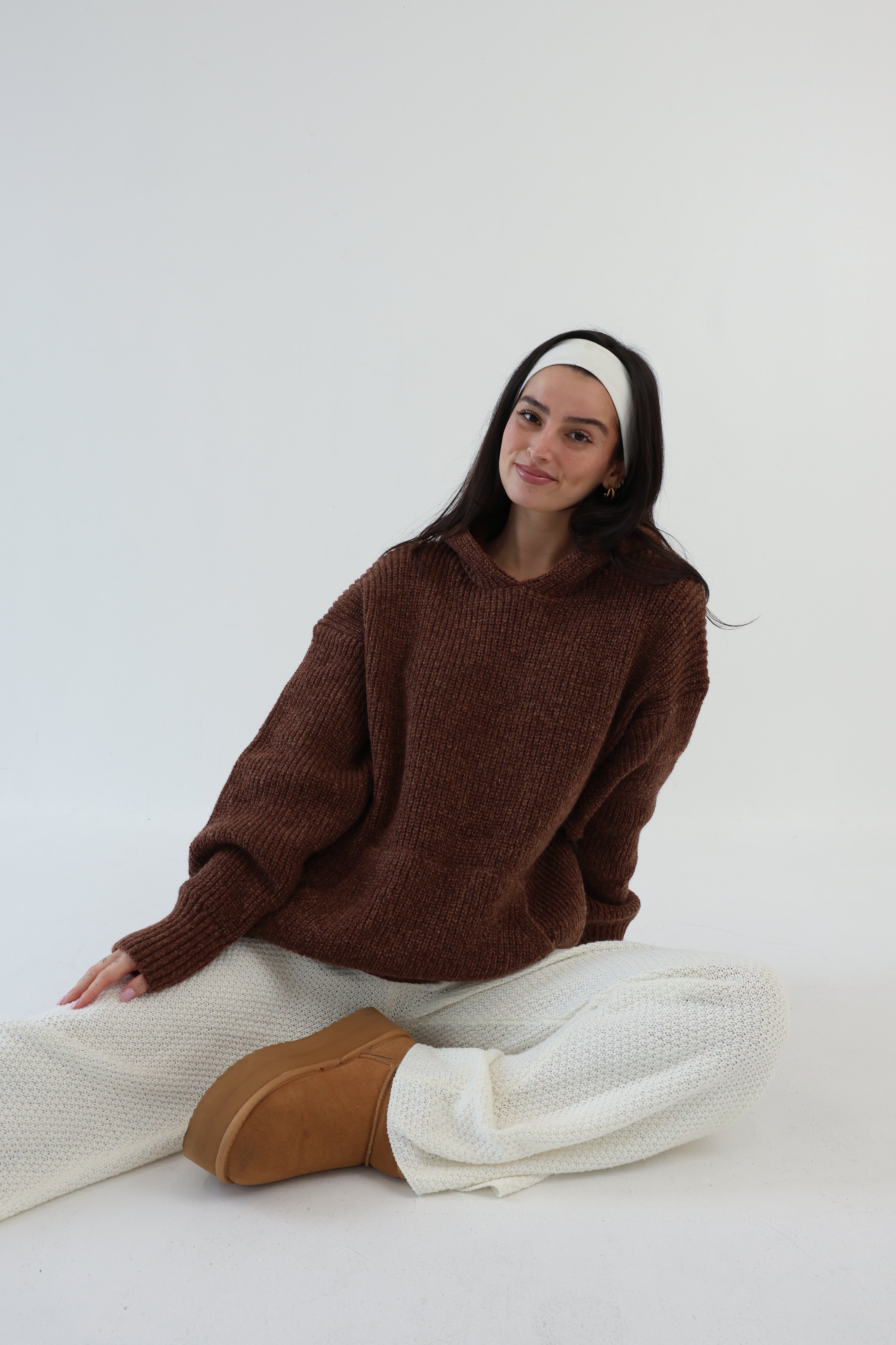 Person wearing a brown sweater and white pants sitting on a white background