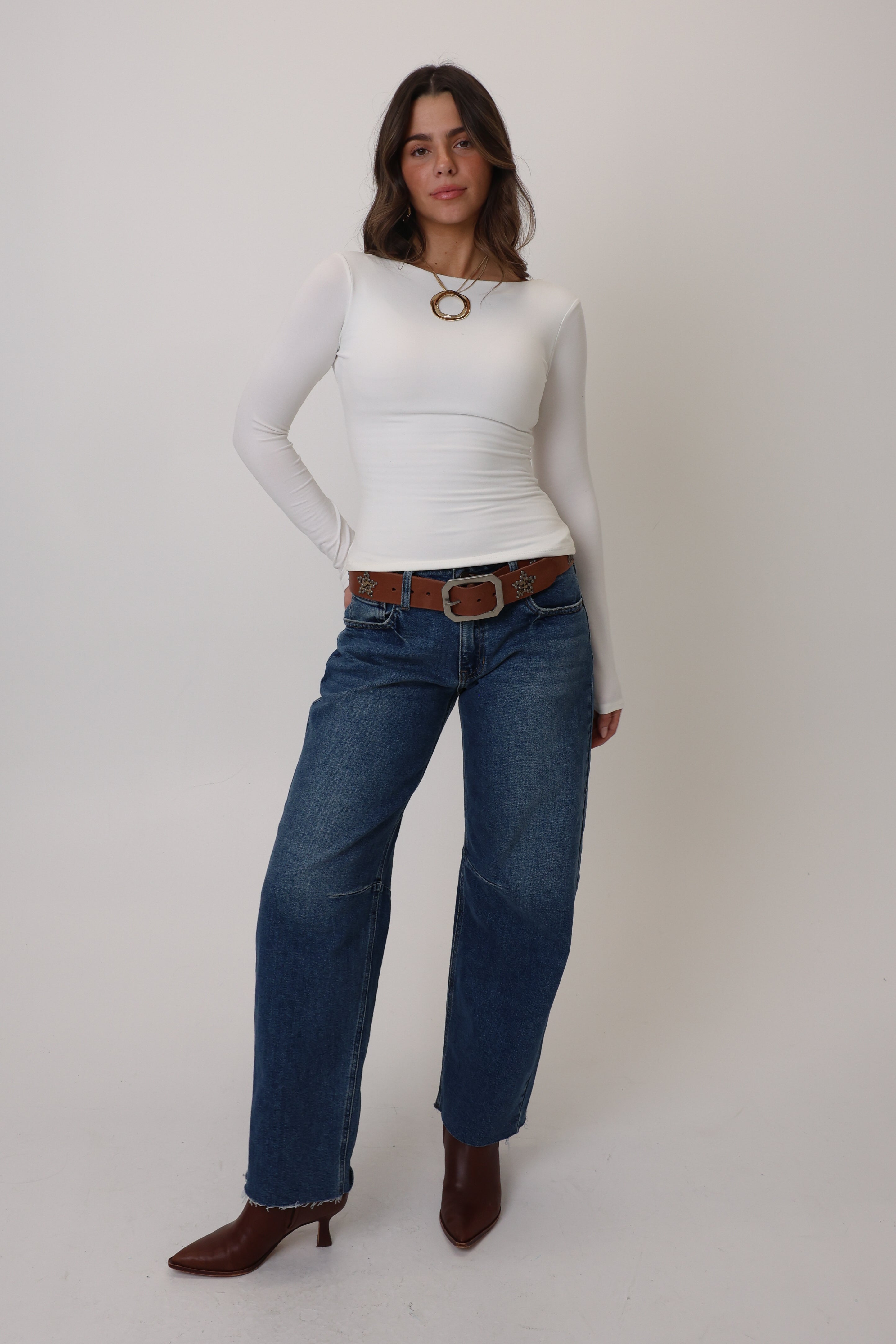 Woman wearing a white long-sleeve top and blue jeans with a brown belt on a white background