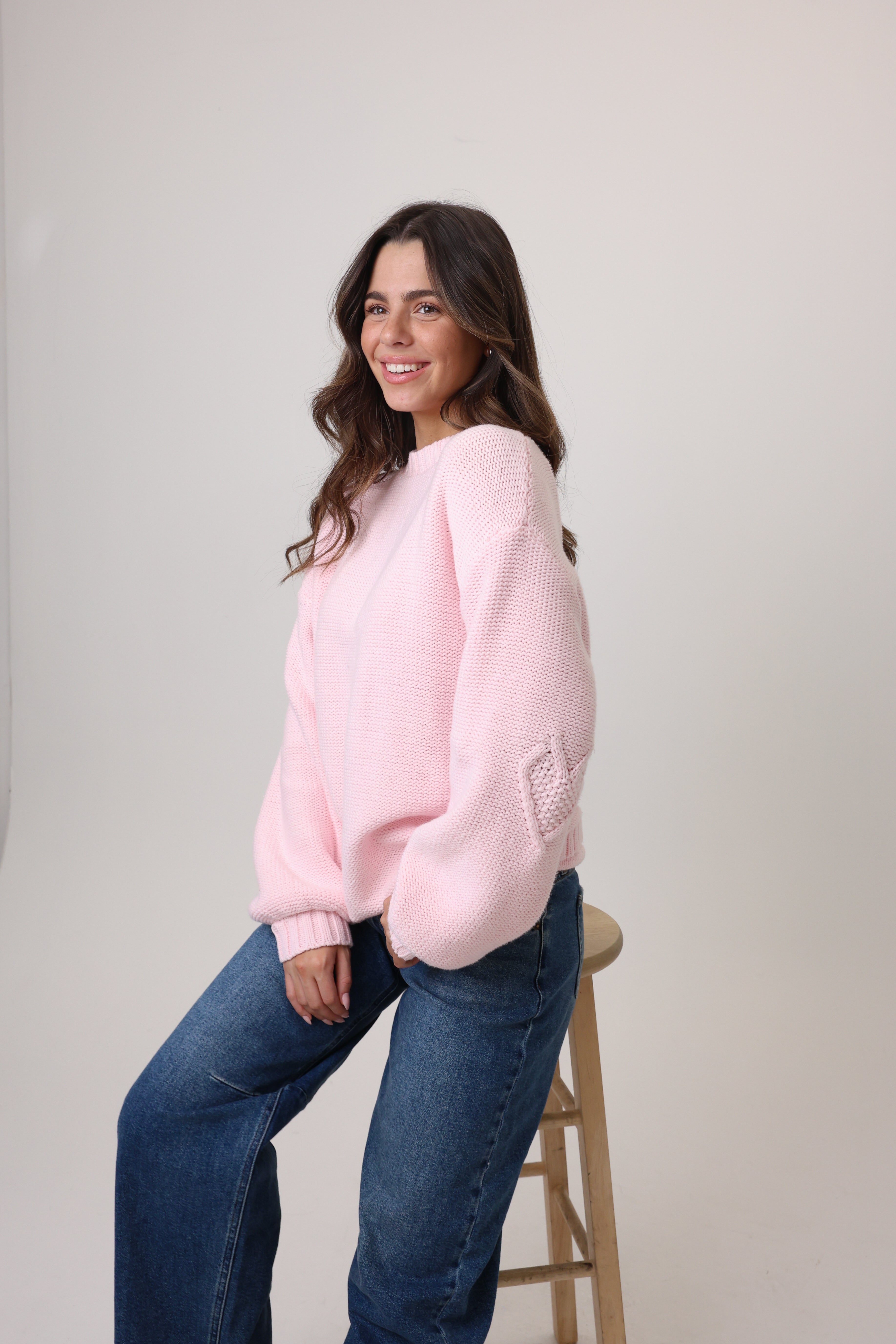 Woman wearing a pink sweater and blue jeans sitting on a wooden stool against a plain background