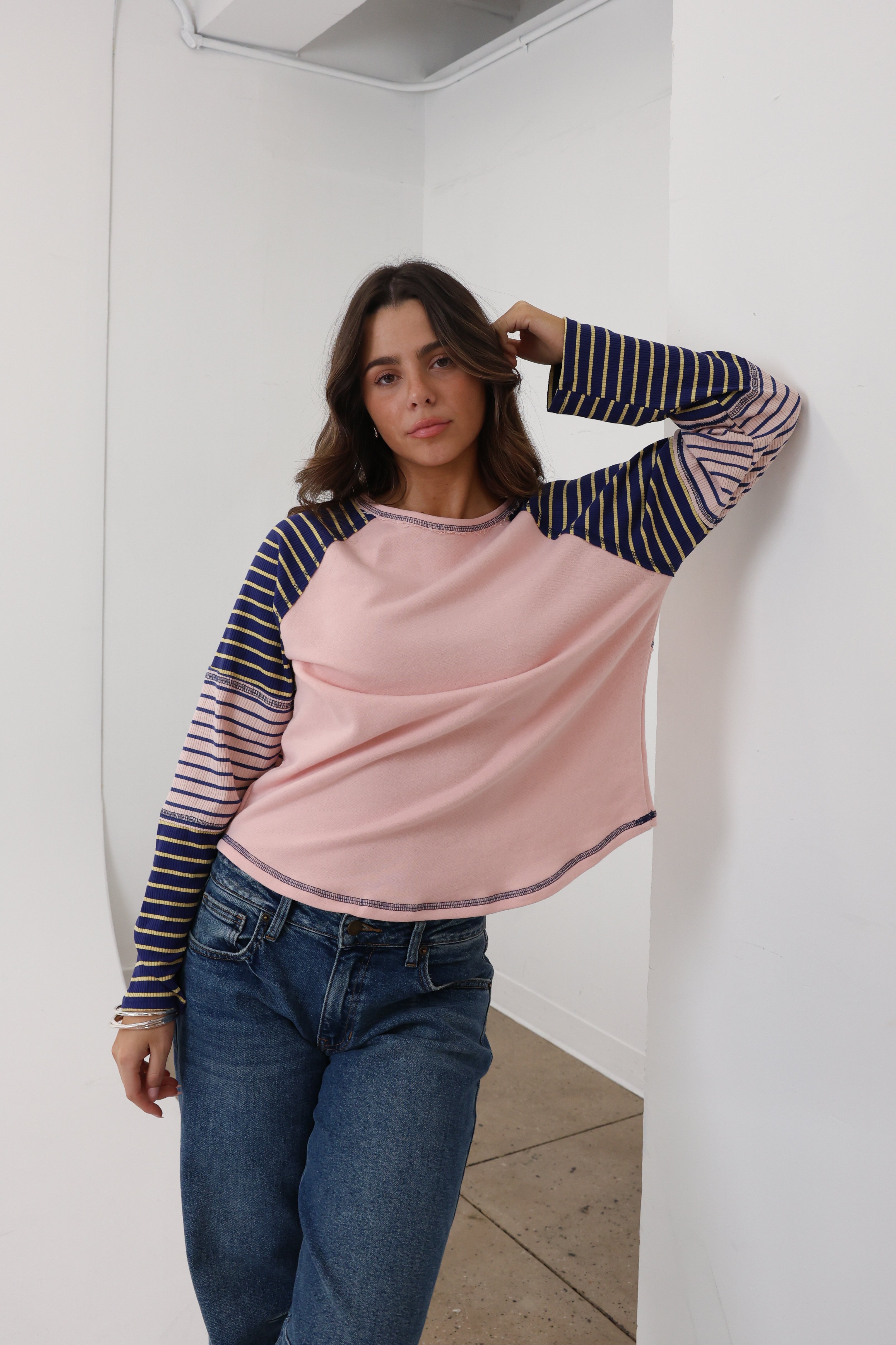 Woman wearing a pink and navy striped long-sleeve top and blue jeans against a white wall.