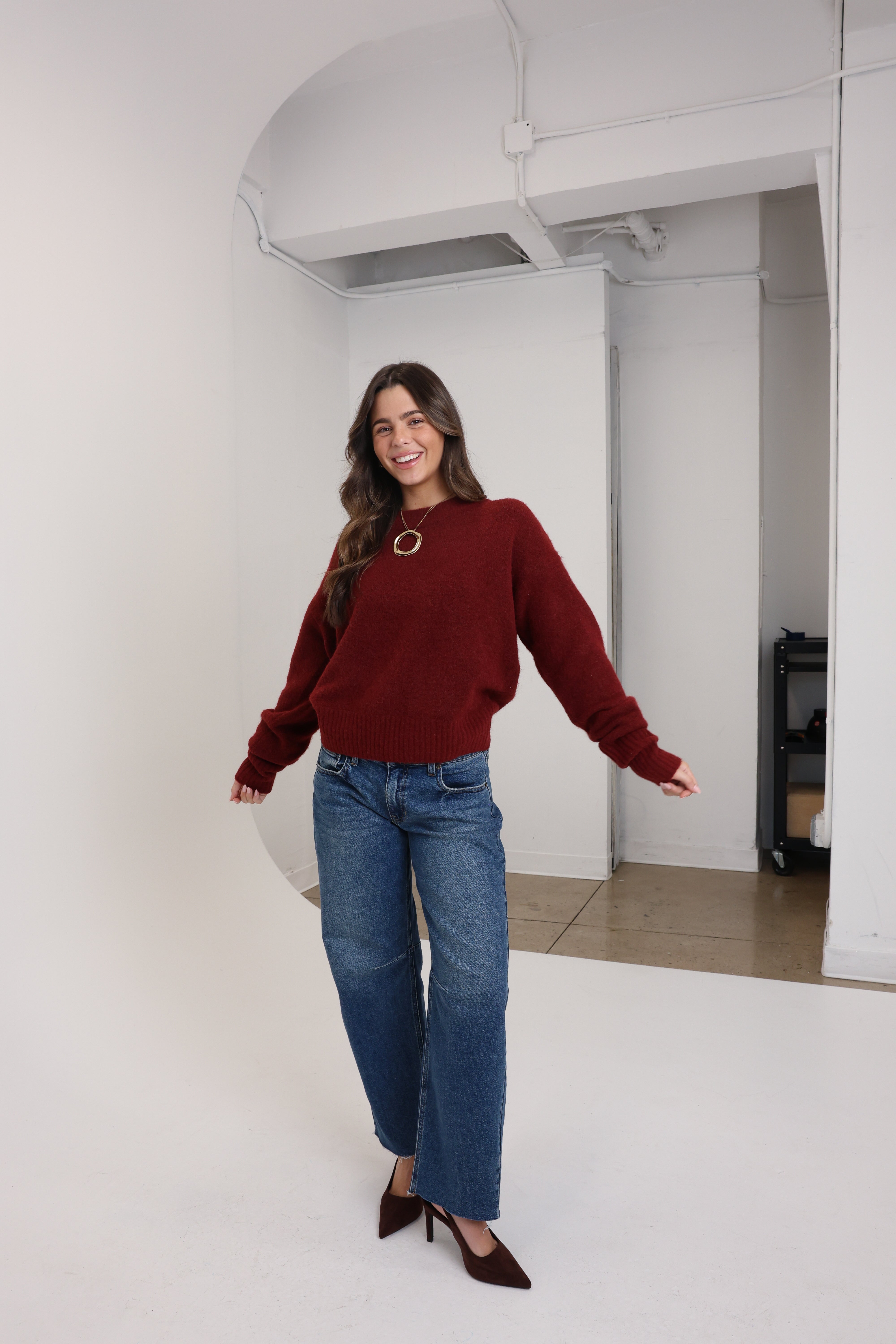 Woman wearing a red sweater and blue jeans standing in a room with white walls.
