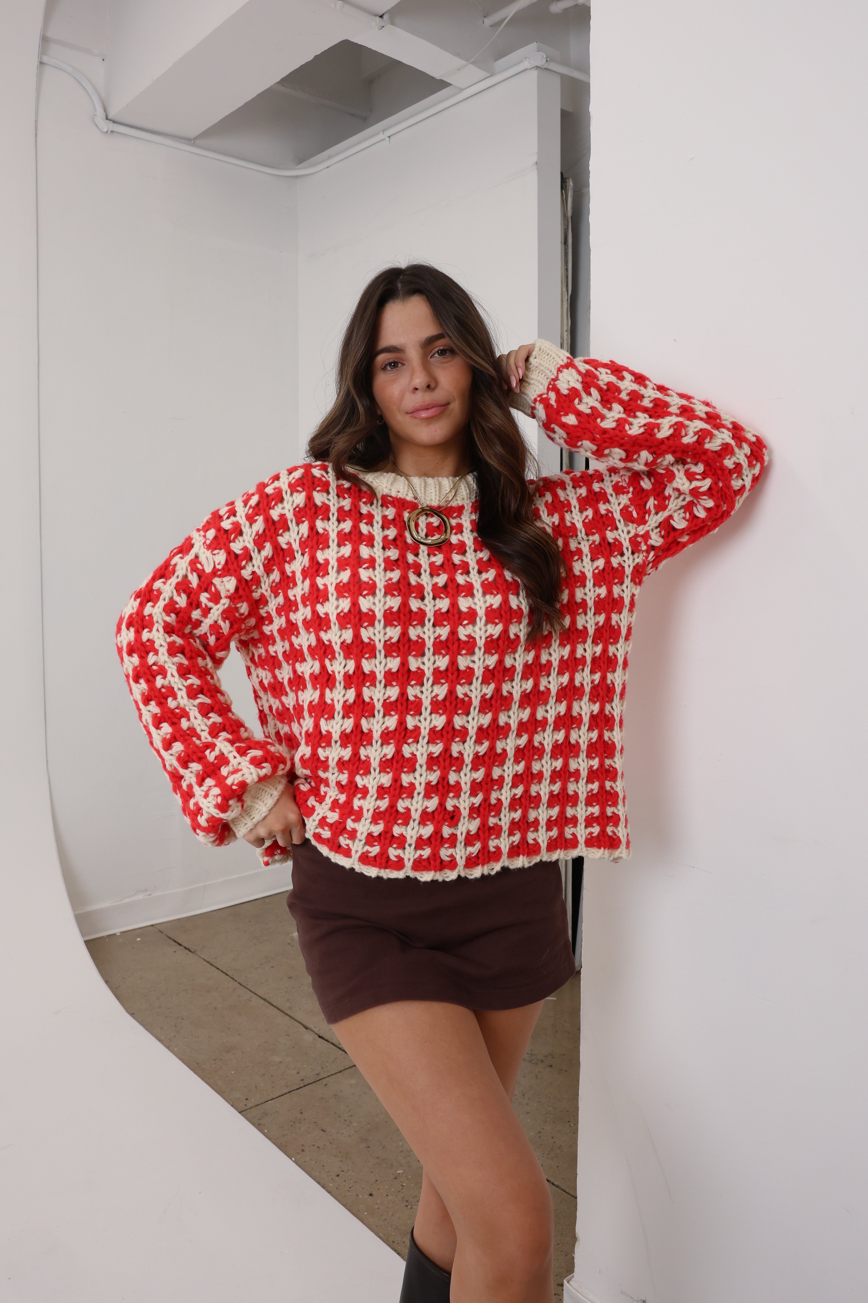 Woman wearing a red and white patterned sweater and brown skirt standing in a room.