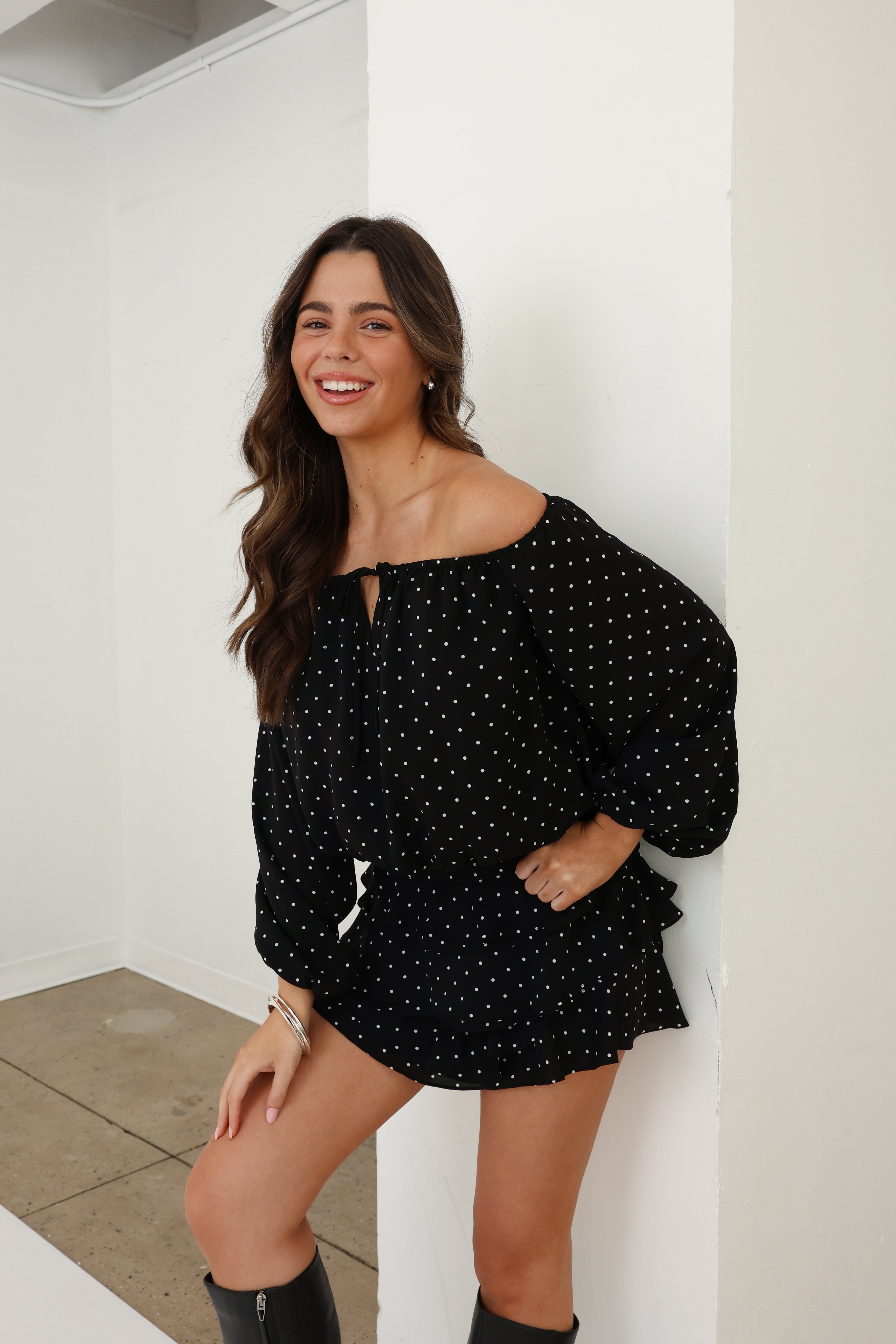 Woman wearing a black polka dot top and shorts, standing against a white wall.
