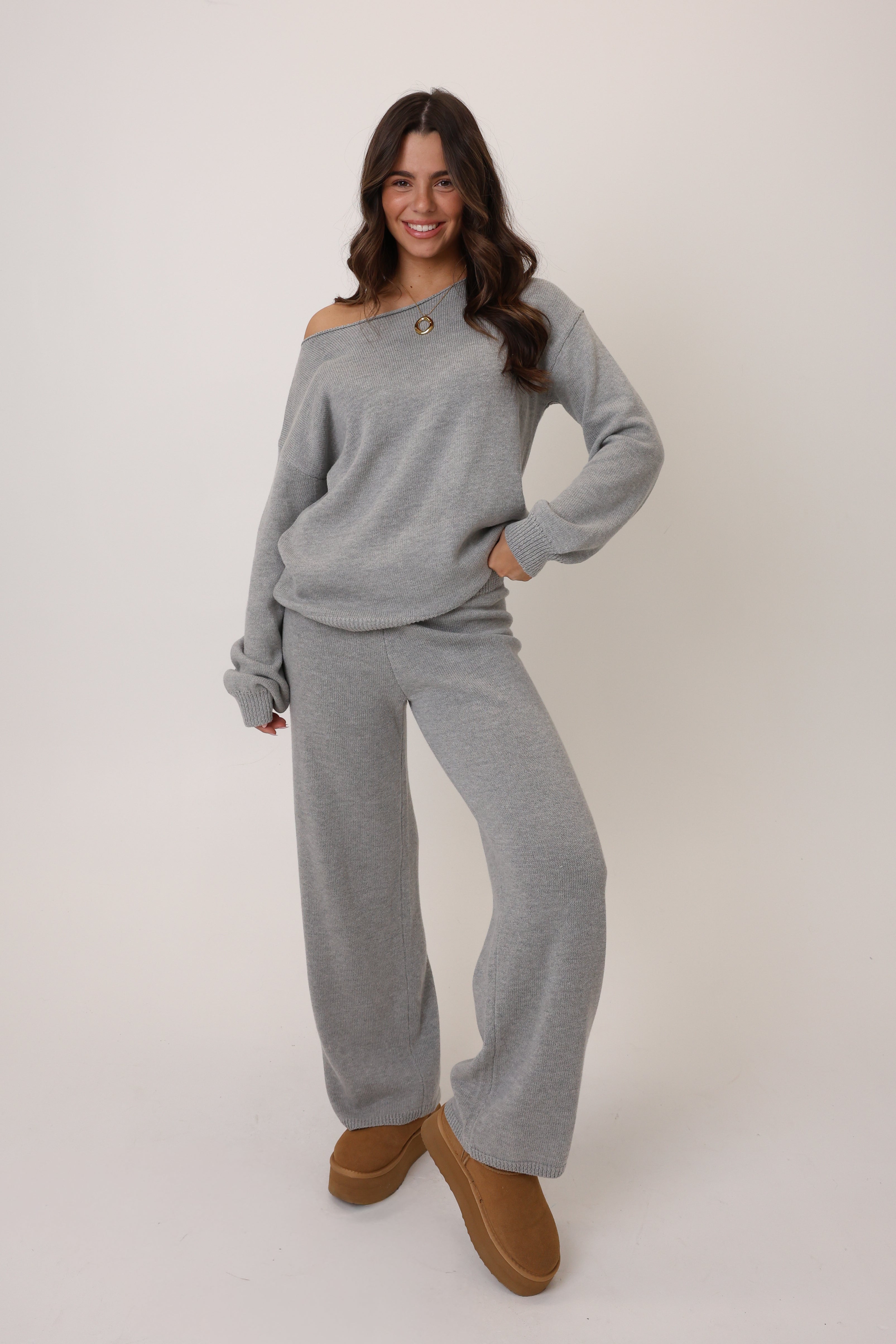 Woman wearing a gray off-shoulder sweater and matching pants with brown boots on a white background