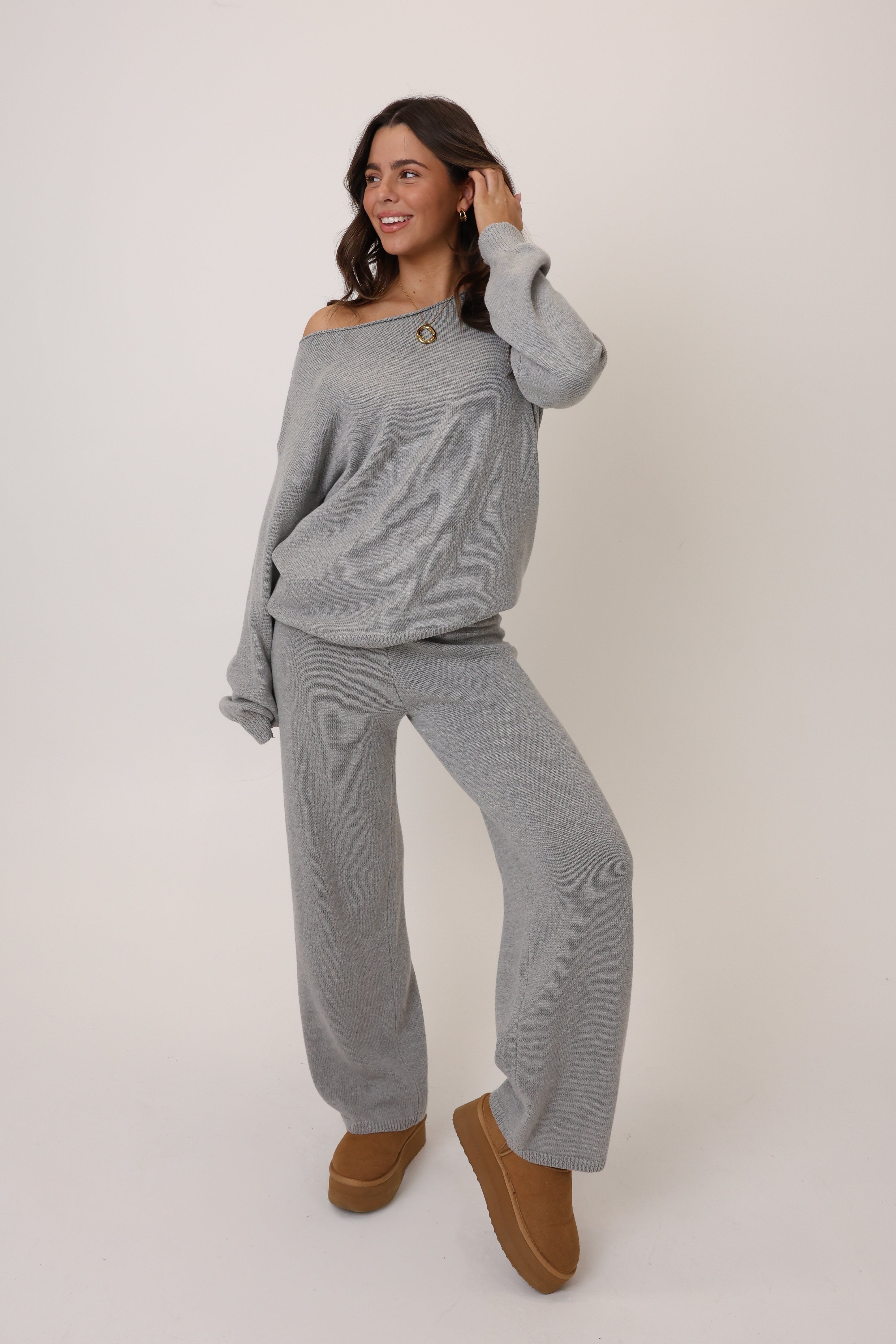 Woman wearing a gray off-shoulder sweater and matching pants with brown shoes on a white background