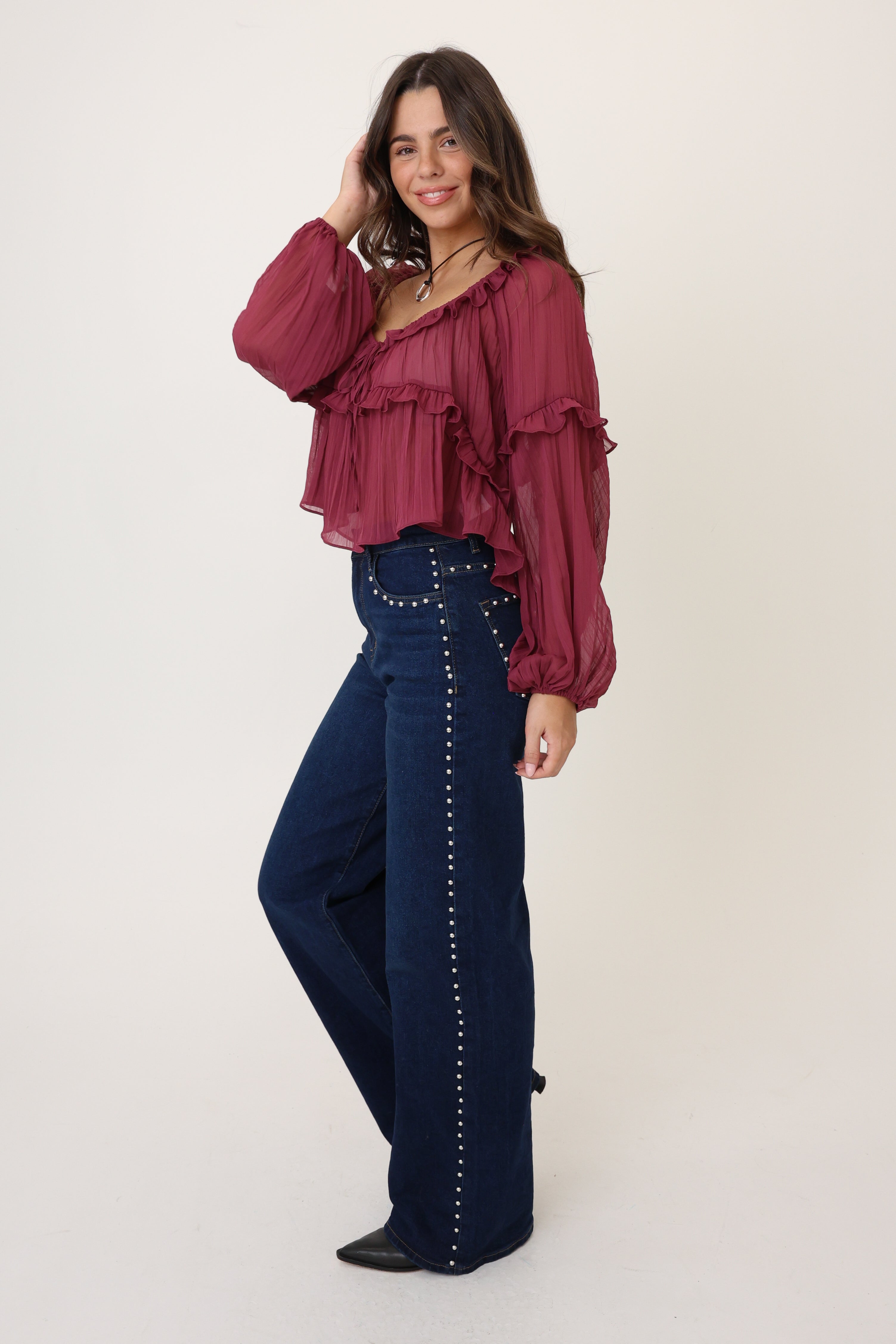 Woman wearing a burgundy blouse and dark blue jeans with studded details on a white background