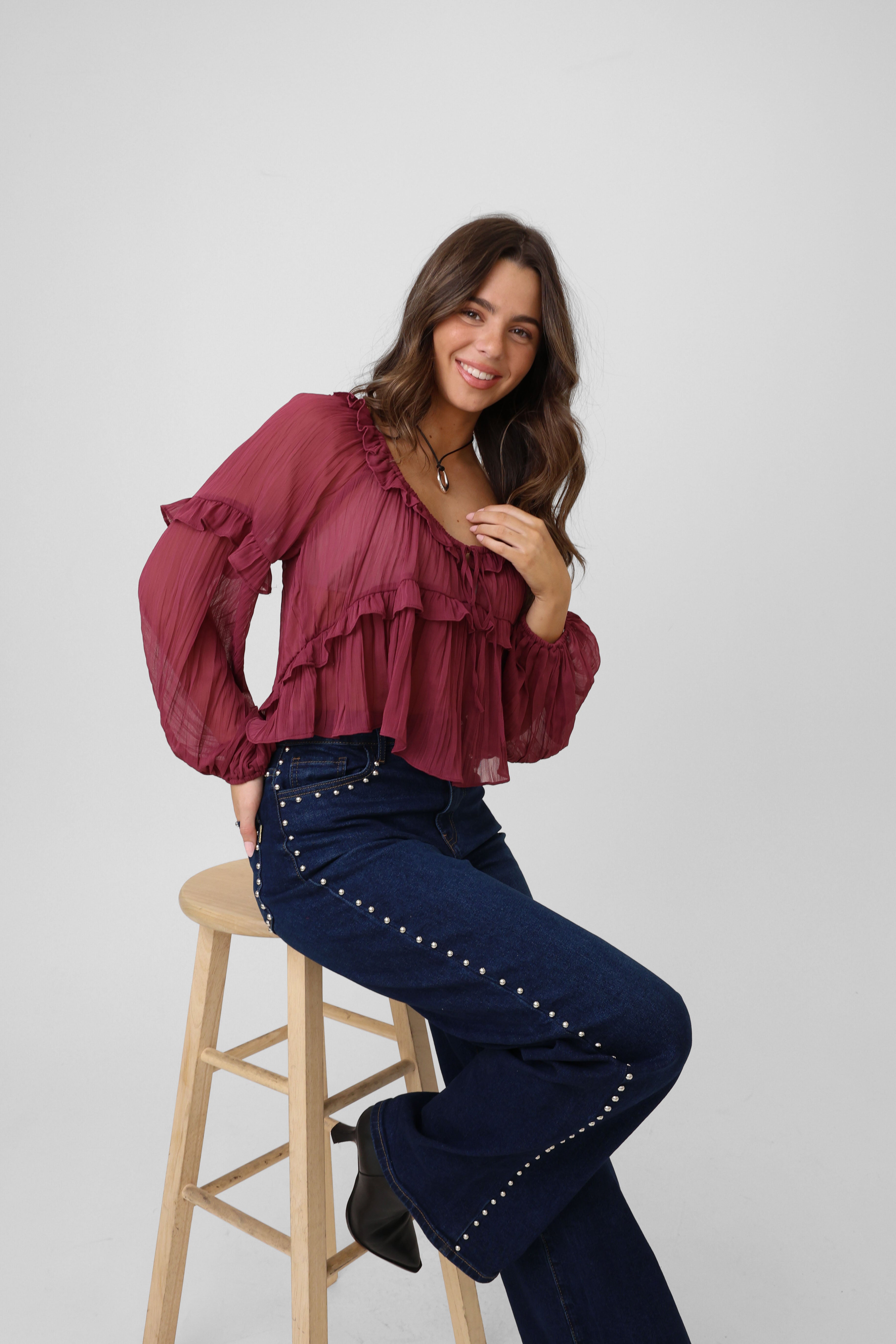 Woman wearing a burgundy blouse and blue pants sitting on a wooden stool against a white background