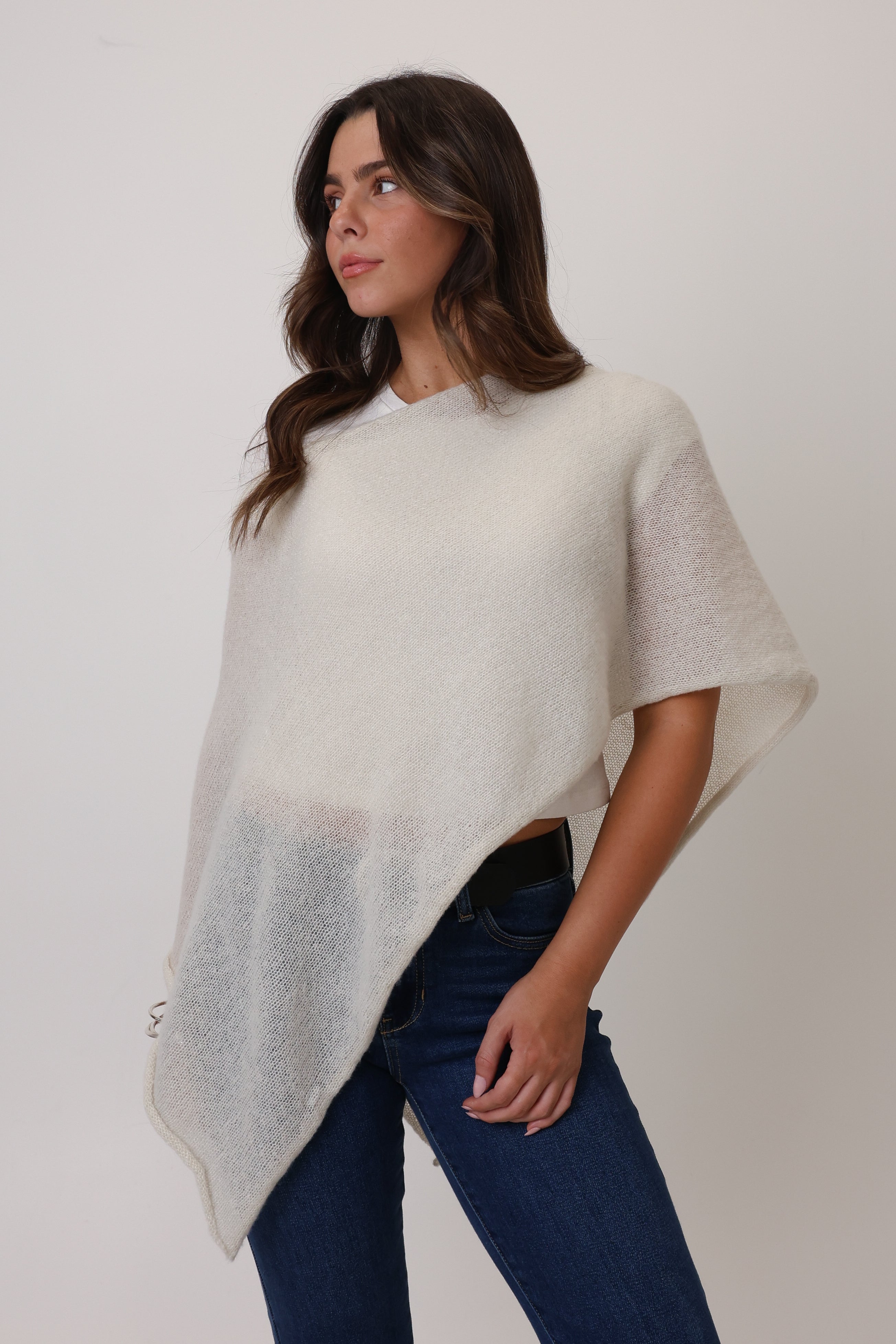 Woman wearing a light gray poncho over a black top and blue jeans on a plain background