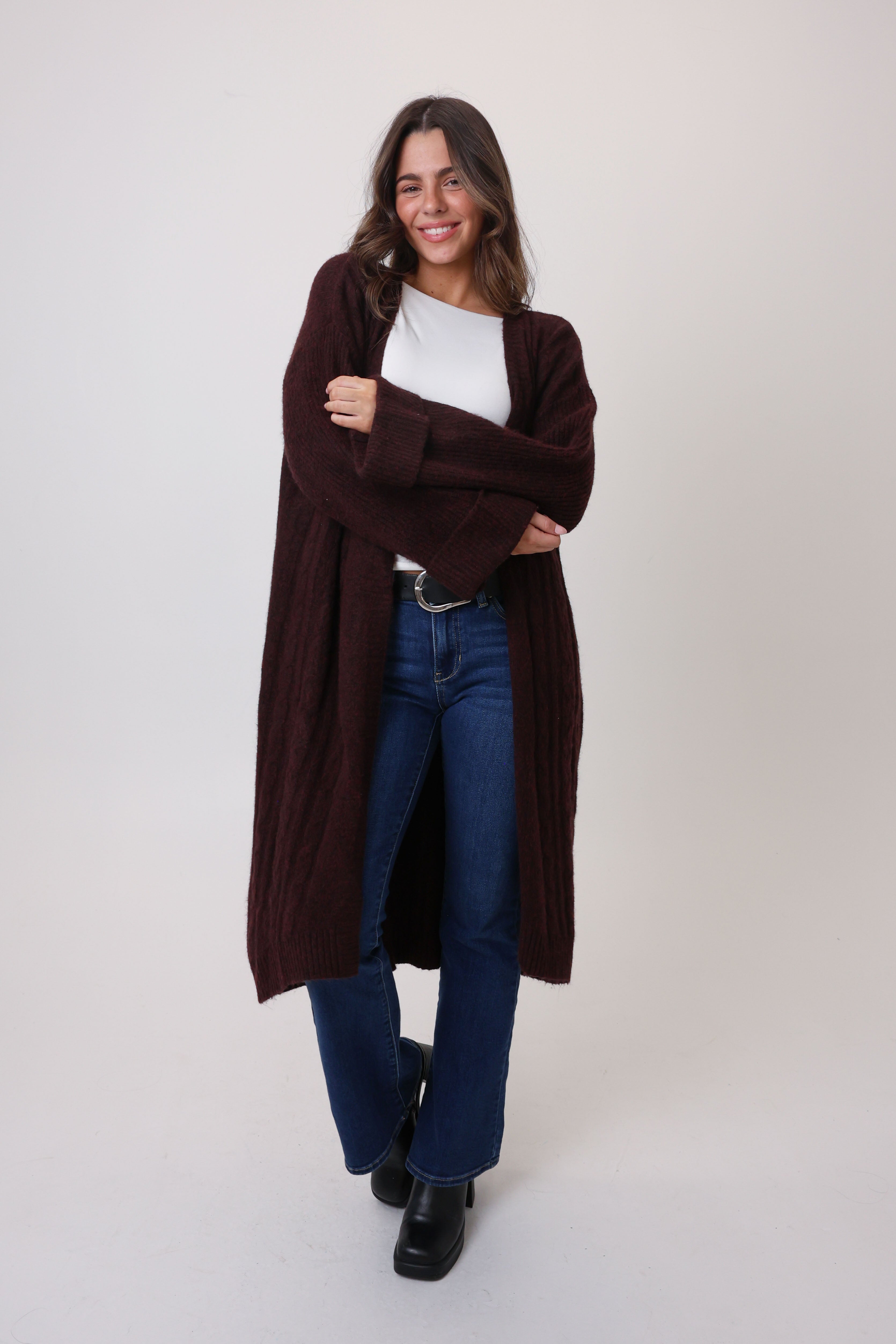 Woman wearing a long brown cardigan over a white shirt and blue jeans on a white background