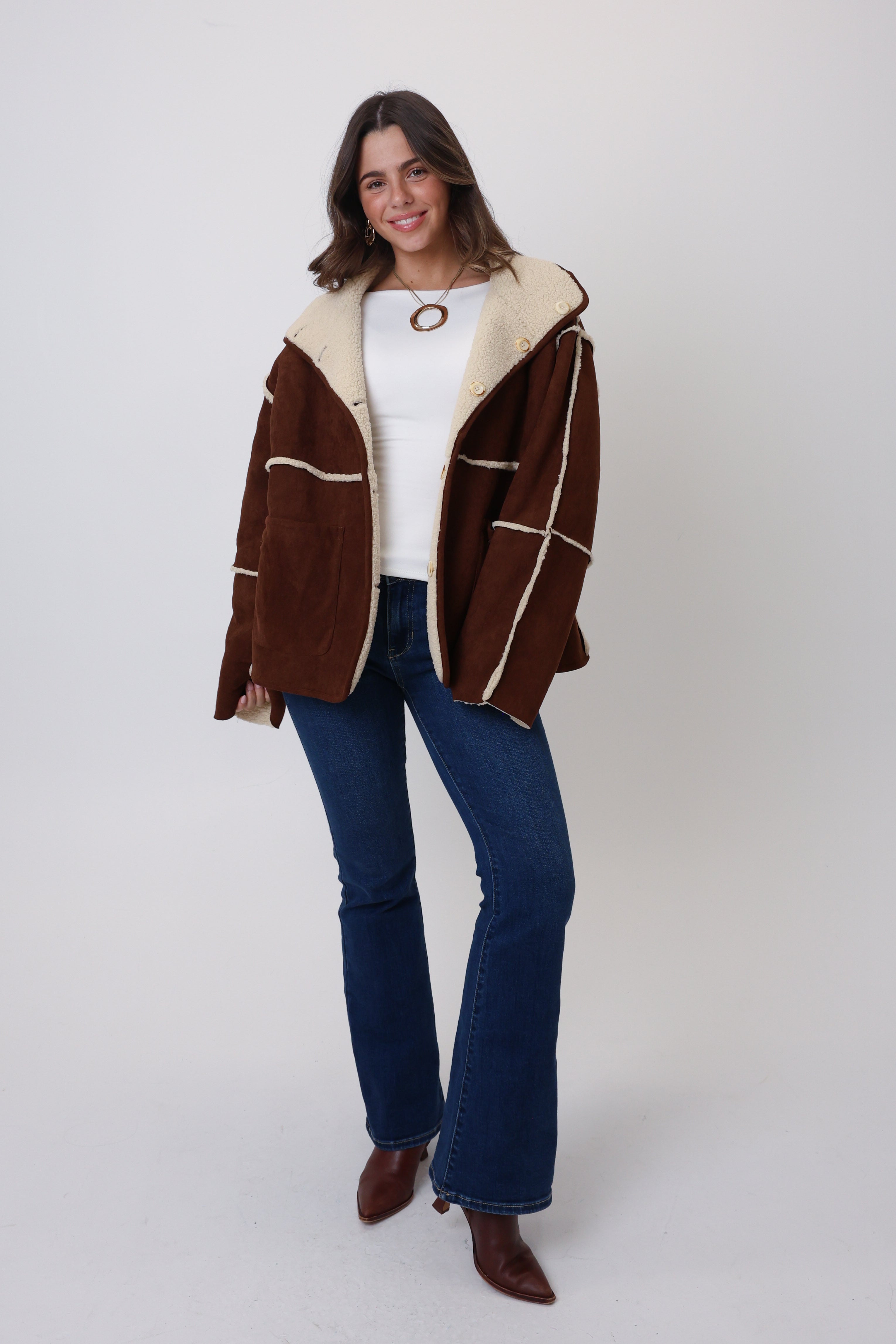 Woman wearing a brown jacket with white fur lining, white shirt, blue jeans, and brown shoes on a white background