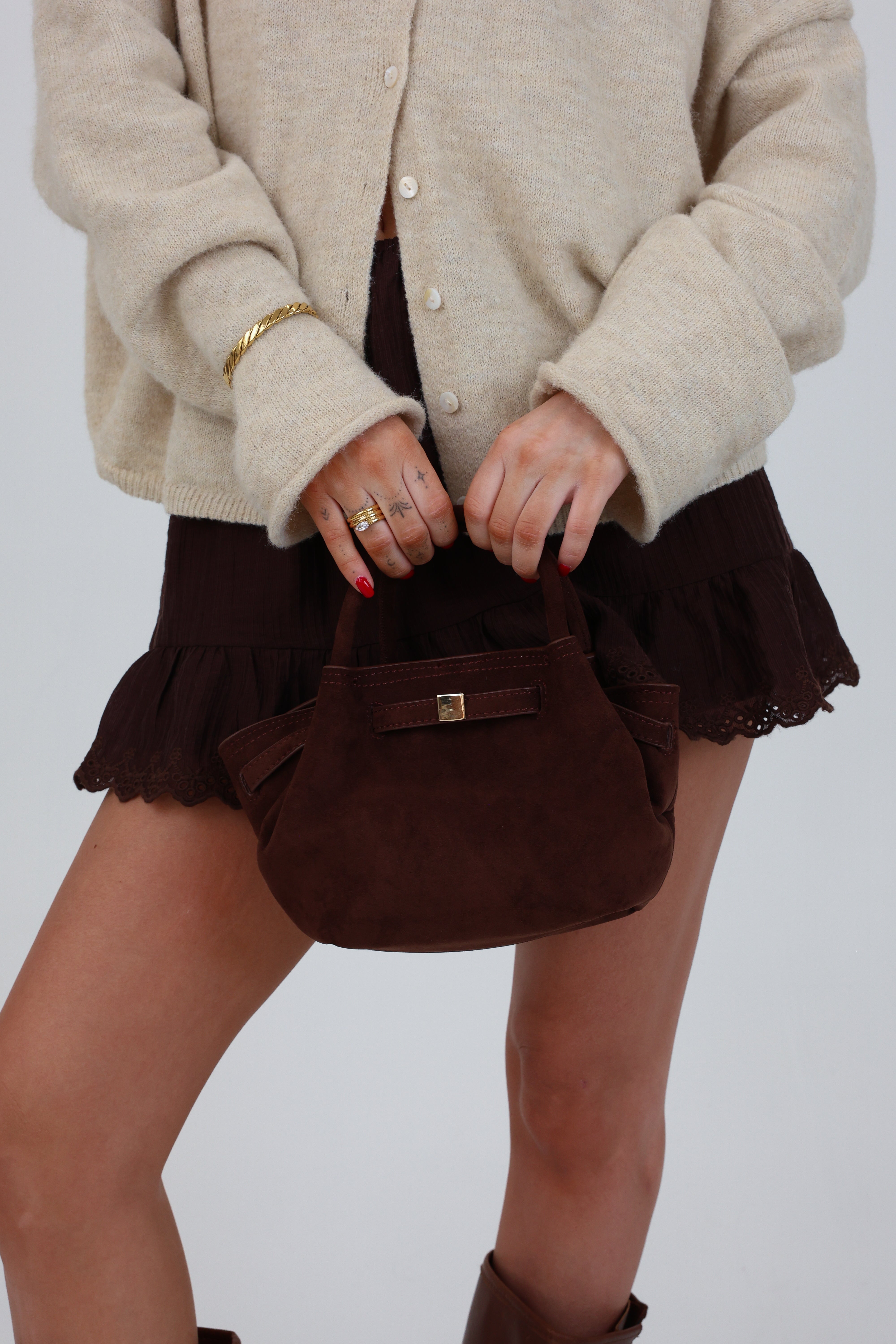 Person wearing a beige cardigan and brown skirt holding a brown handbag against a plain background
