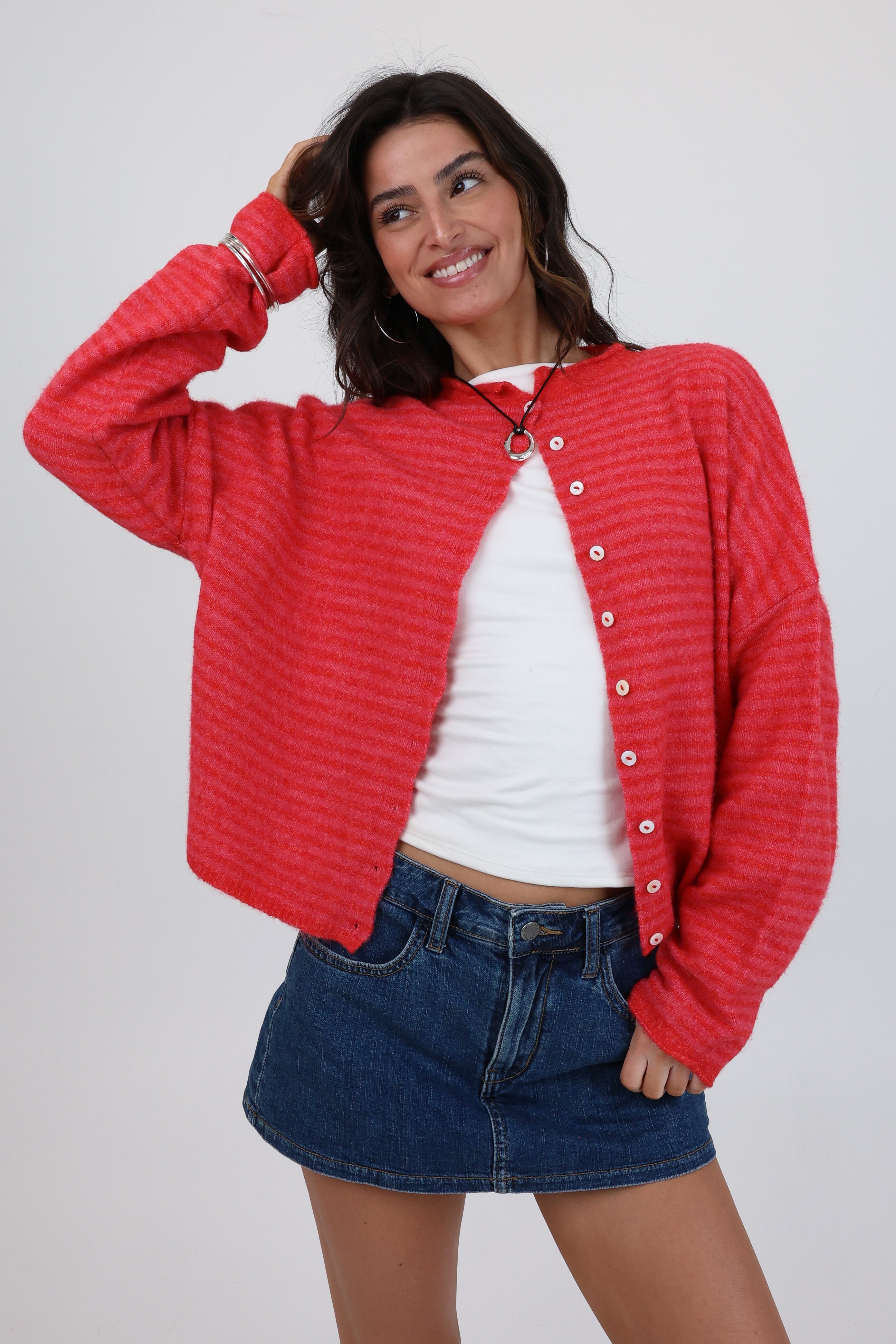 Woman wearing a red textured cardigan over a white top and blue shorts on a plain background
