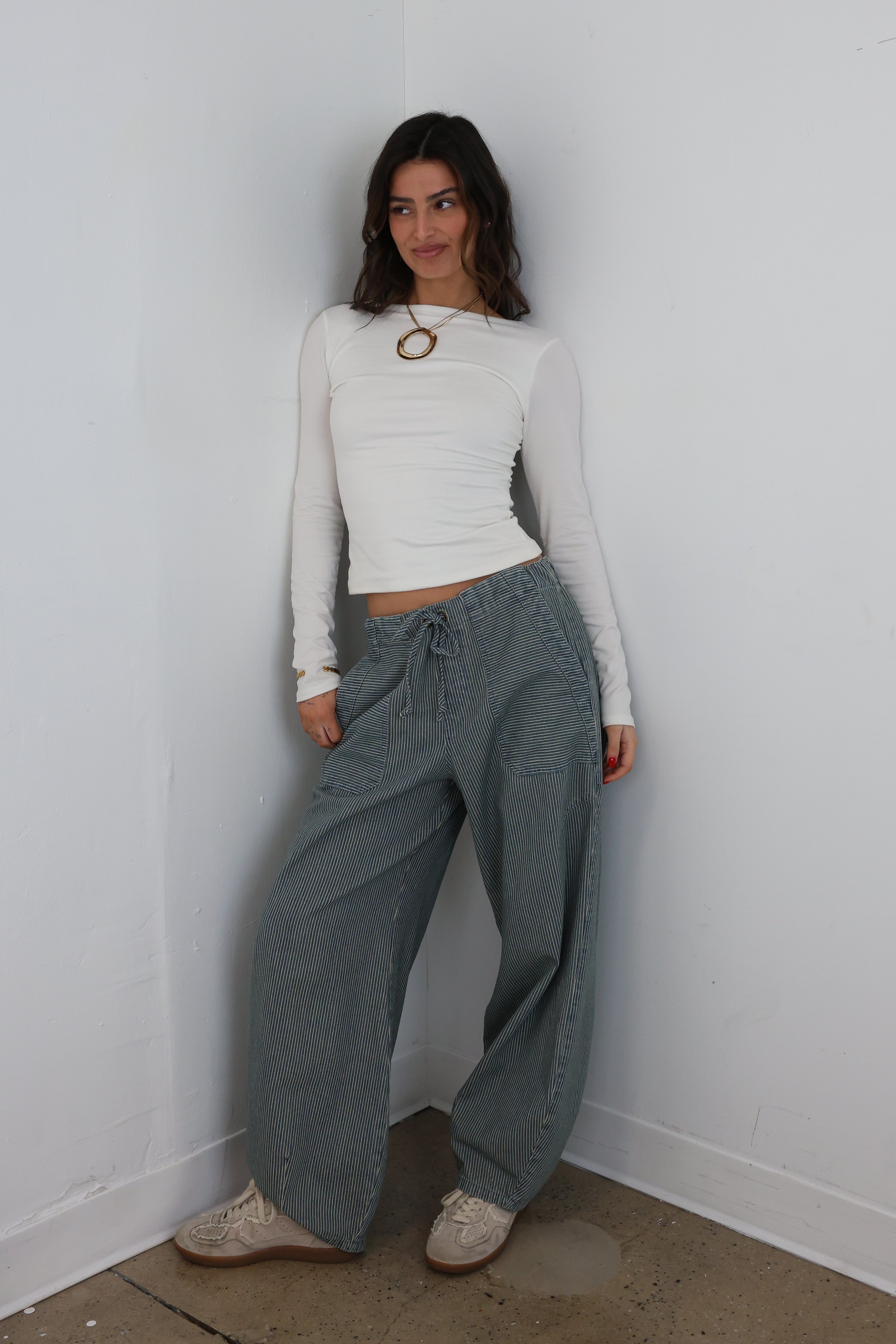 Woman wearing a white long-sleeve top and gray pants standing against a white wall.