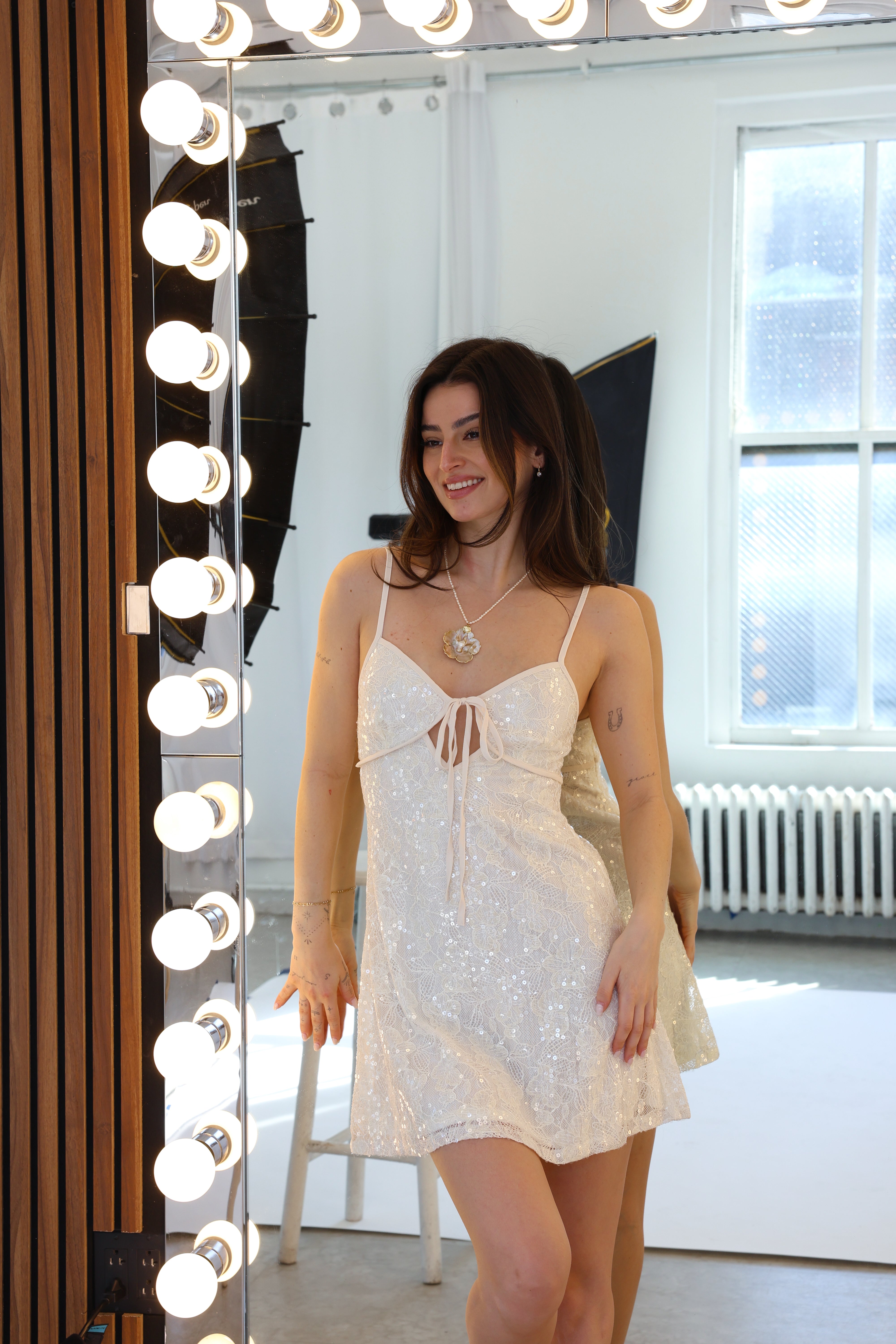 Woman in a white lace dress standing in front of a mirror with lights.