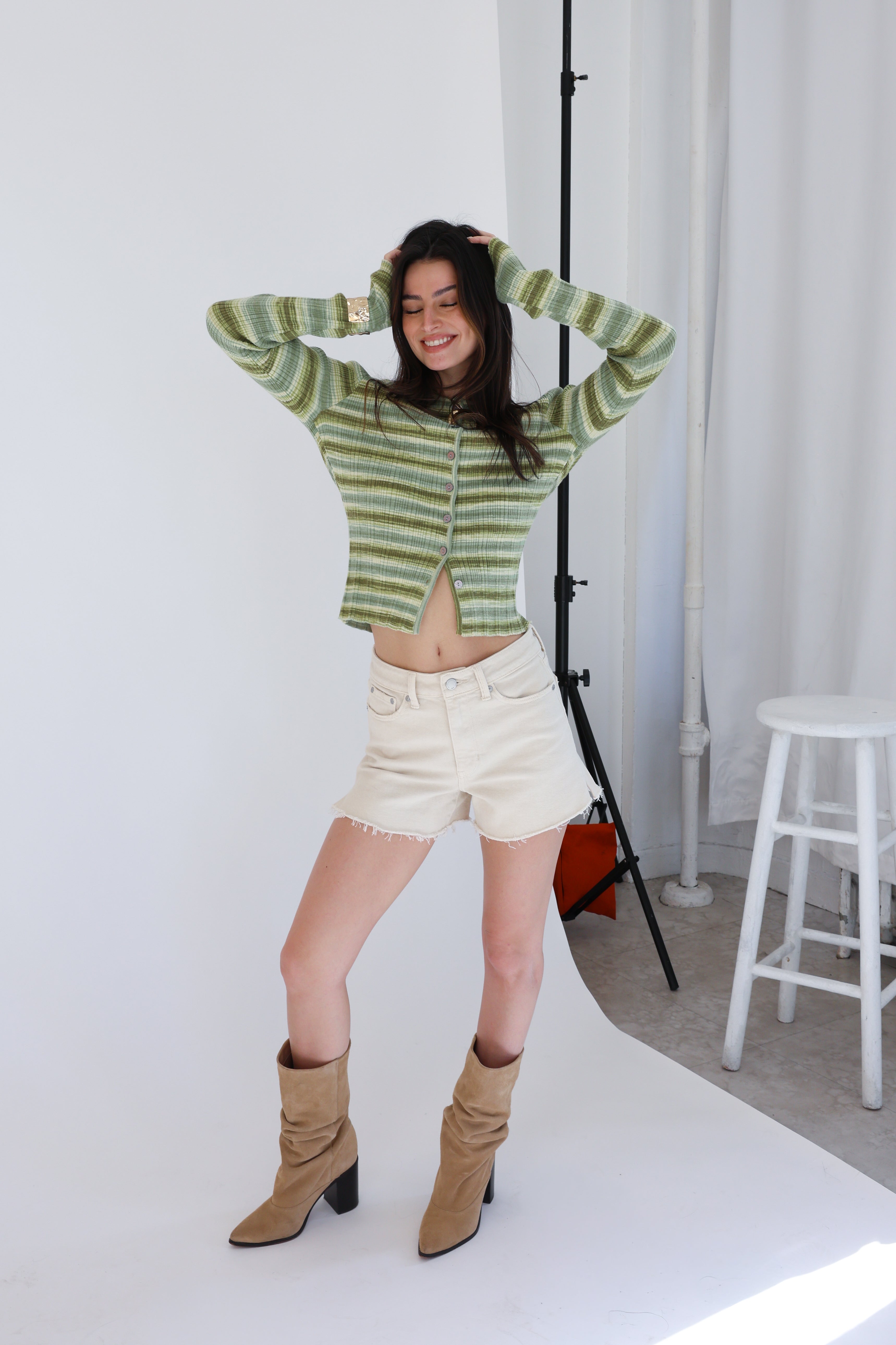 Person wearing a green striped sweater and beige shorts standing in a studio setting.