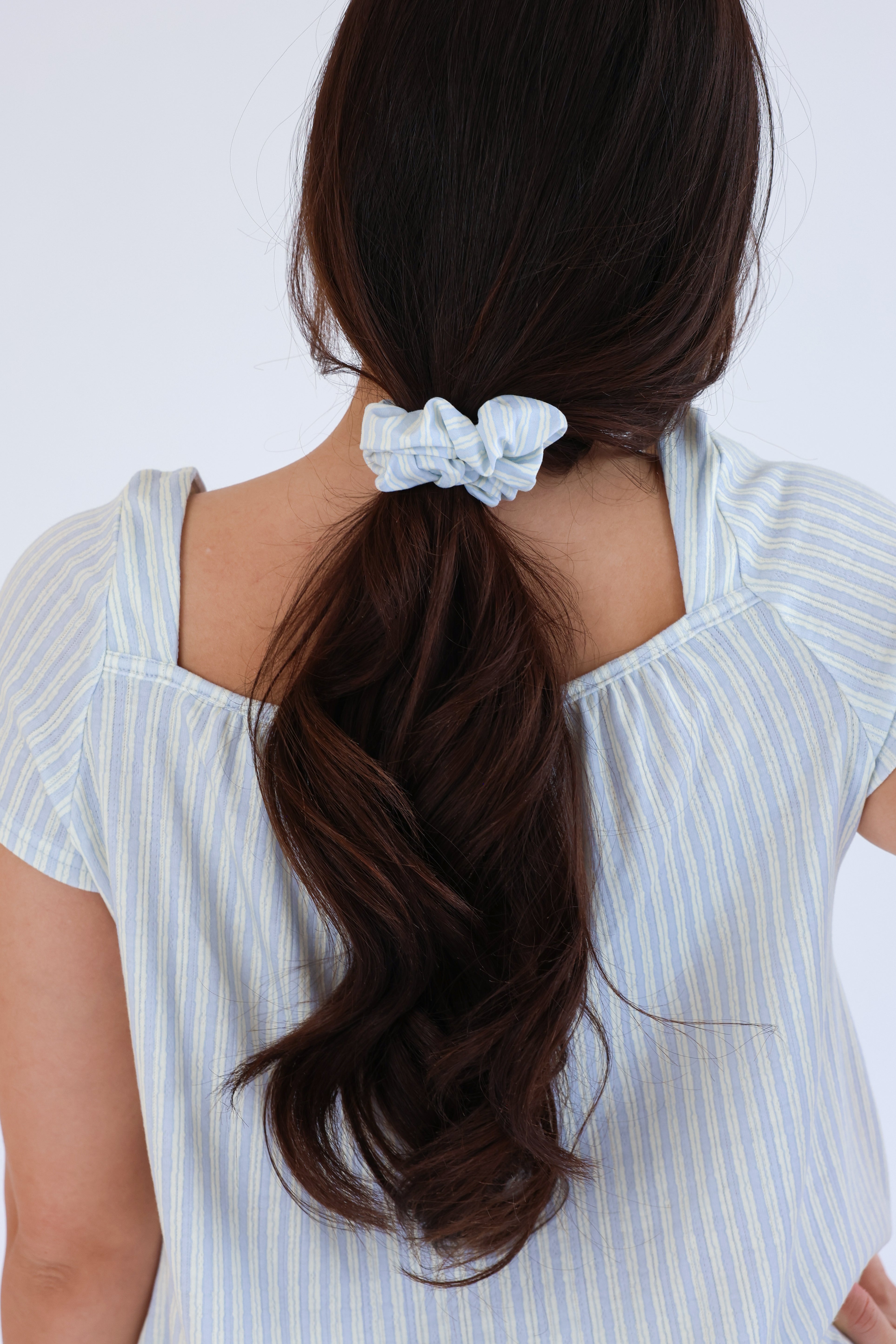 Woman with long dark hair tied back with a light blue scrunchie against a white background