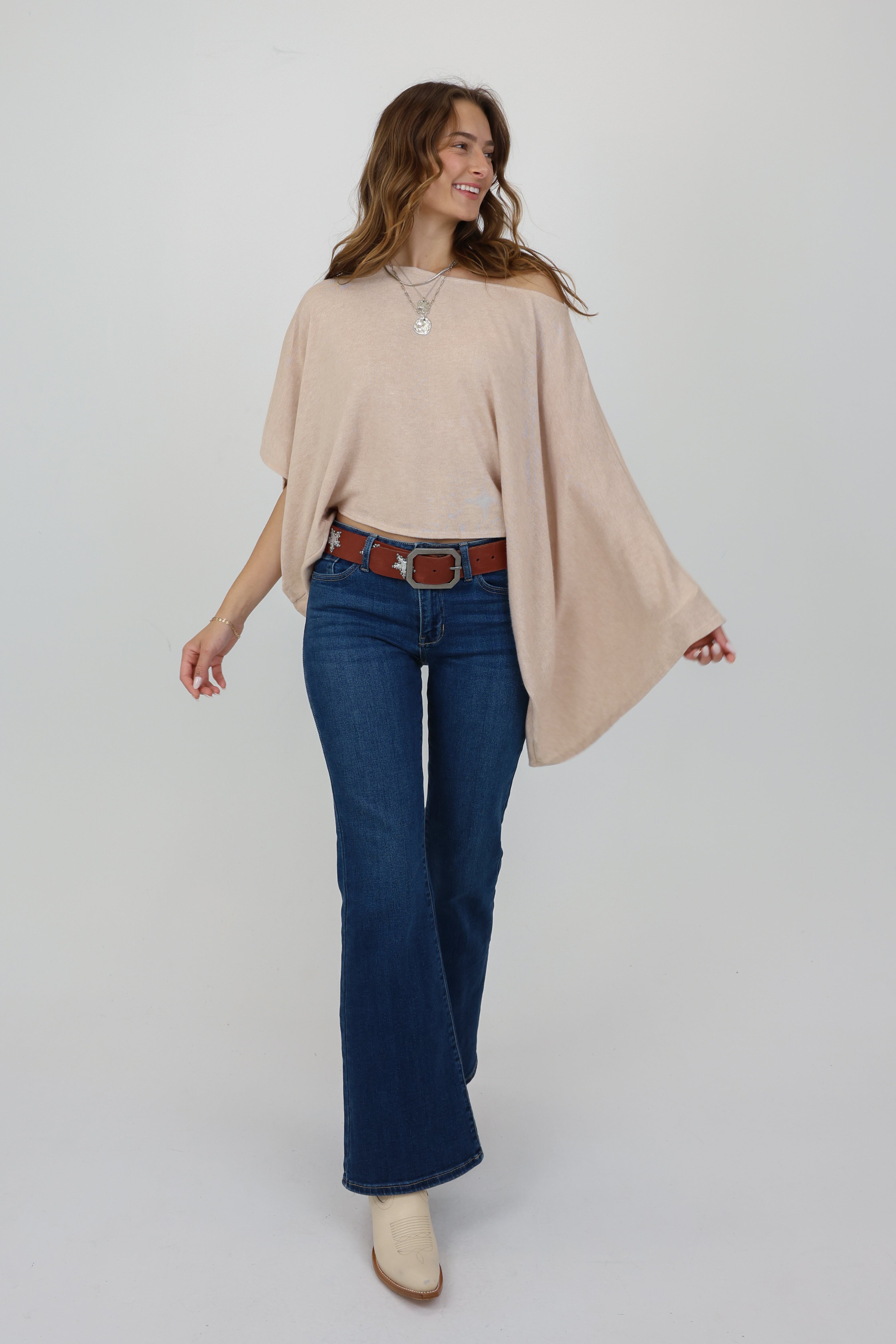 Woman wearing a beige poncho, blue jeans, and white shoes on a plain background