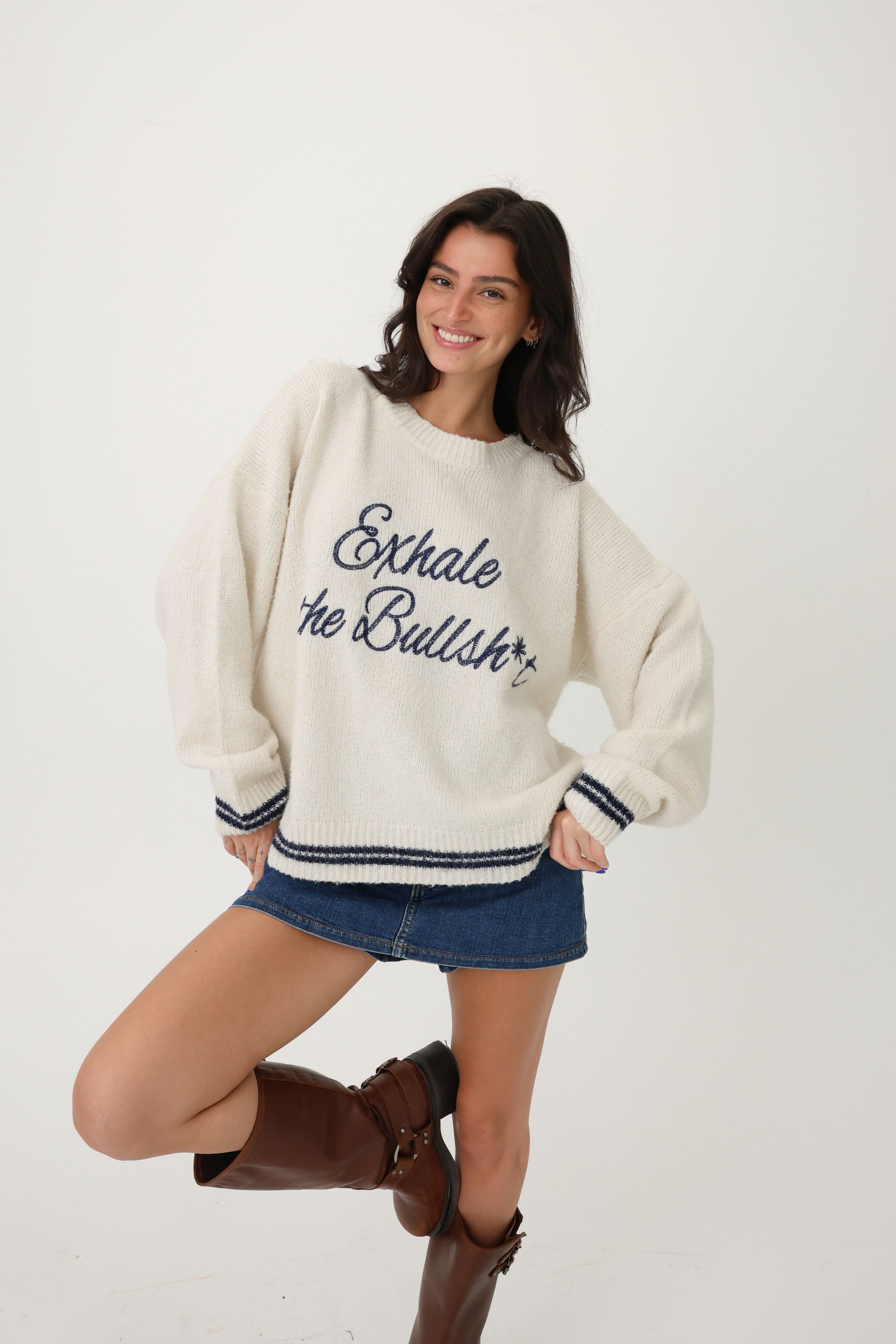 Woman wearing a cream sweater with text, denim shorts, and brown boots on a white background