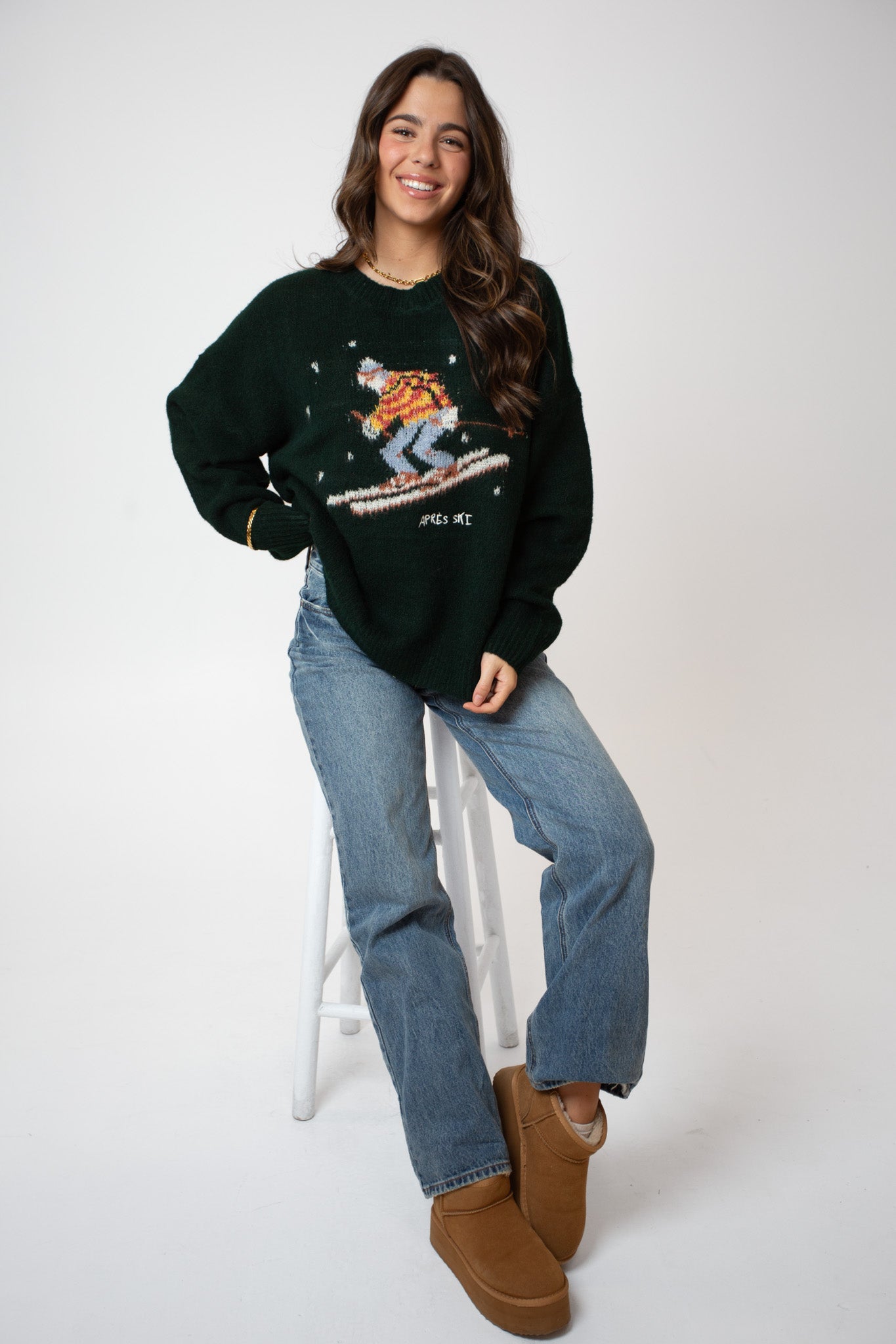 Woman wearing a dark green sweater with a graphic design, blue jeans, and brown shoes on a white background