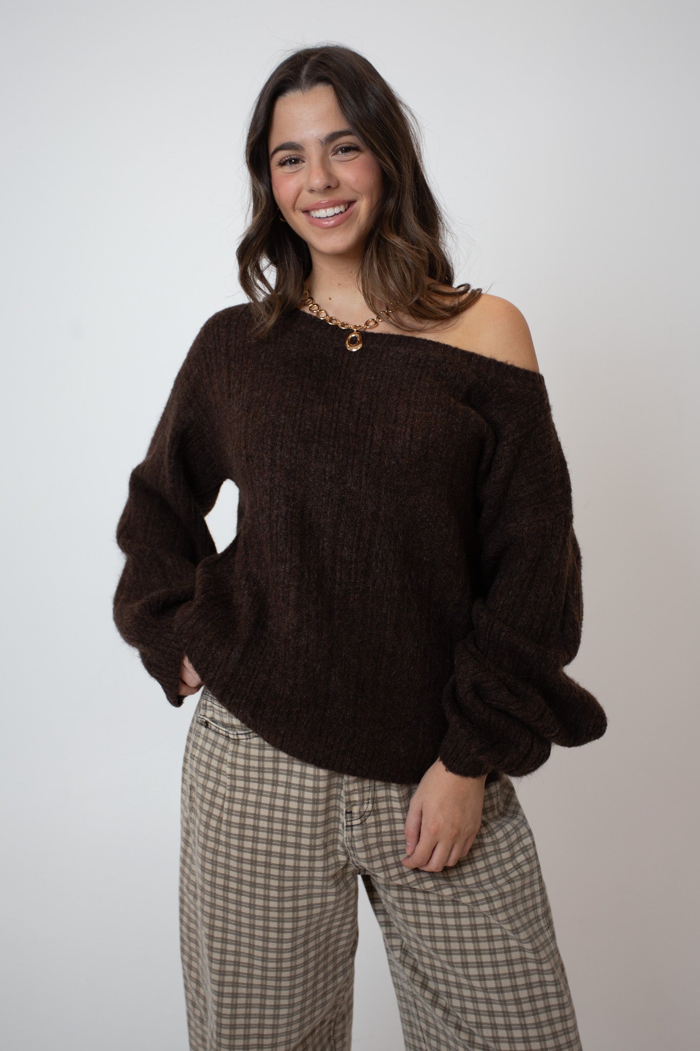 Woman wearing a brown off-shoulder sweater and plaid pants against a plain background