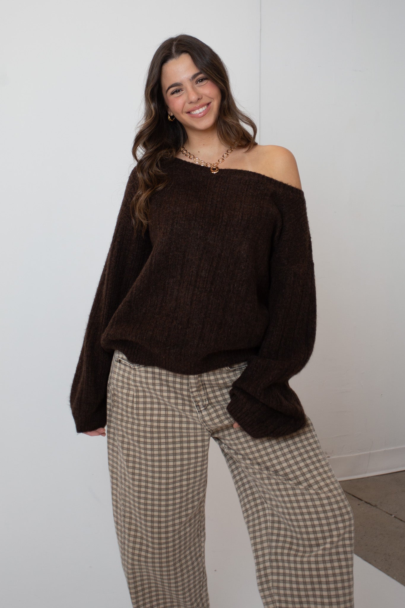 Woman wearing a brown off-shoulder sweater and checkered pants against a white wall.