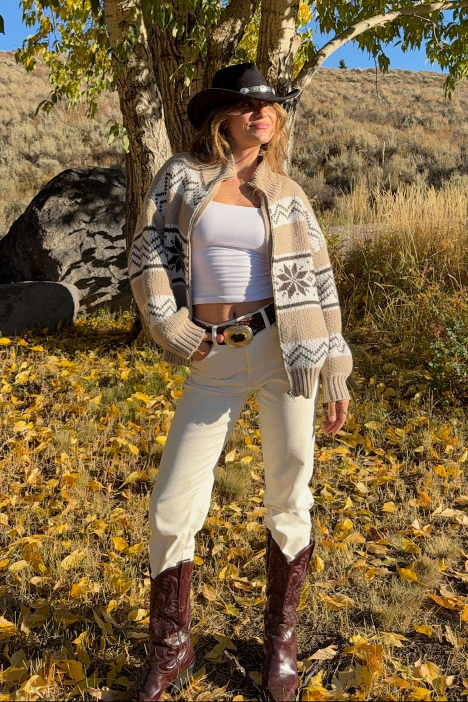 Person wearing a patterned cardigan, white pants, and cowboy boots in a natural setting with trees and grass.