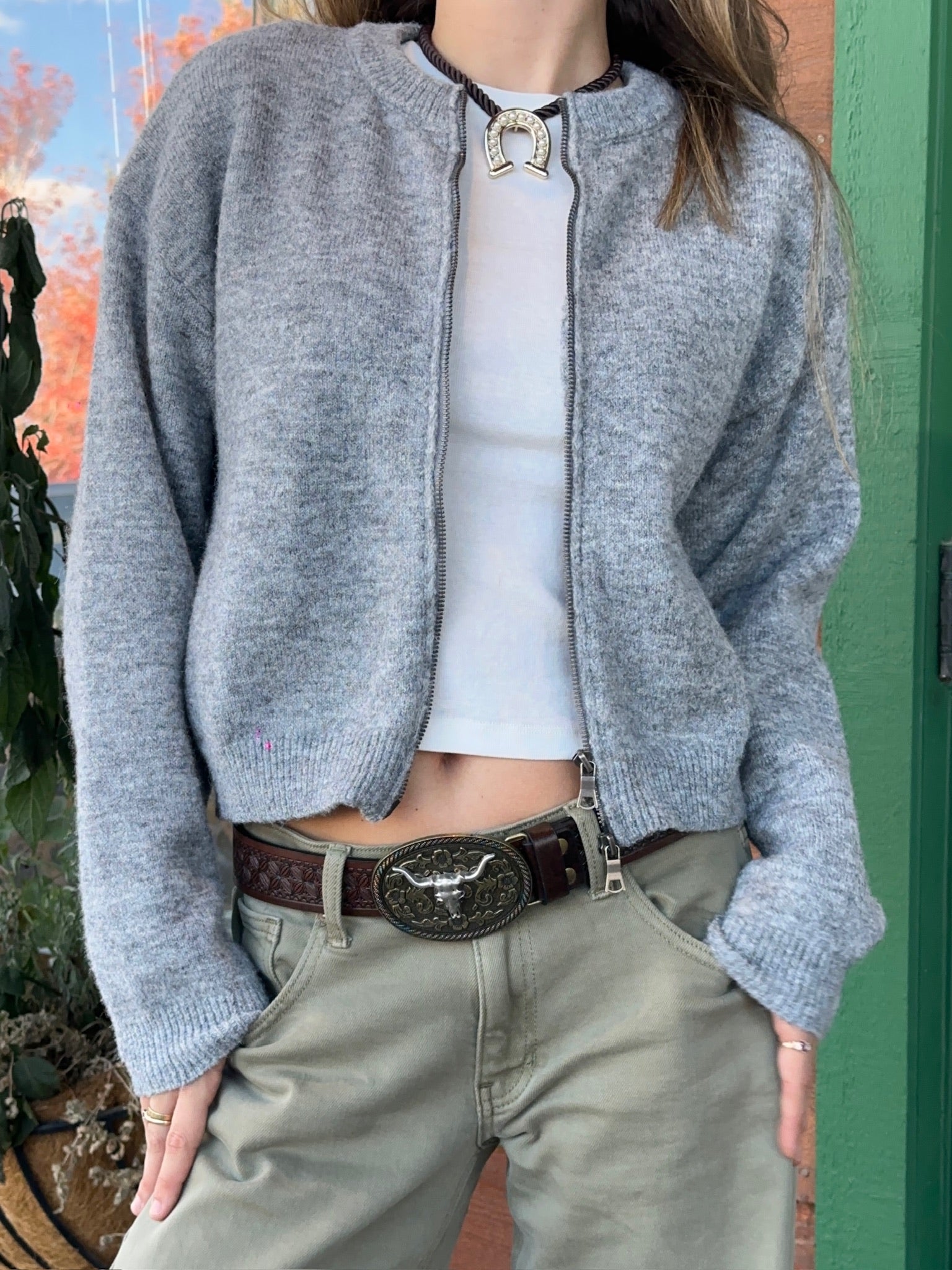 Person wearing a gray cardigan over a white top and khaki pants with a belt, standing outdoors.