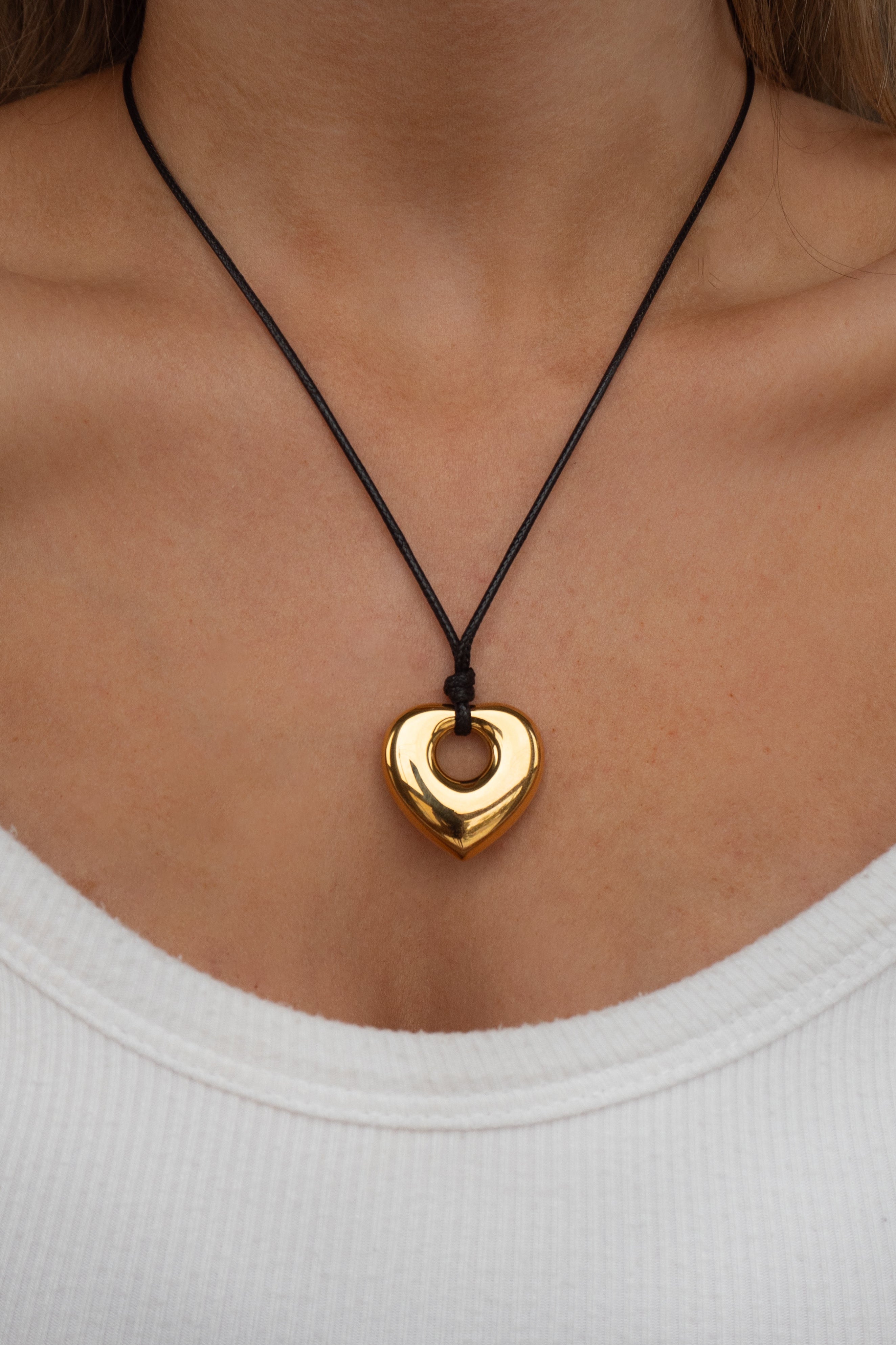 Gold heart-shaped pendant on a black cord necklace worn by a person in a white top.