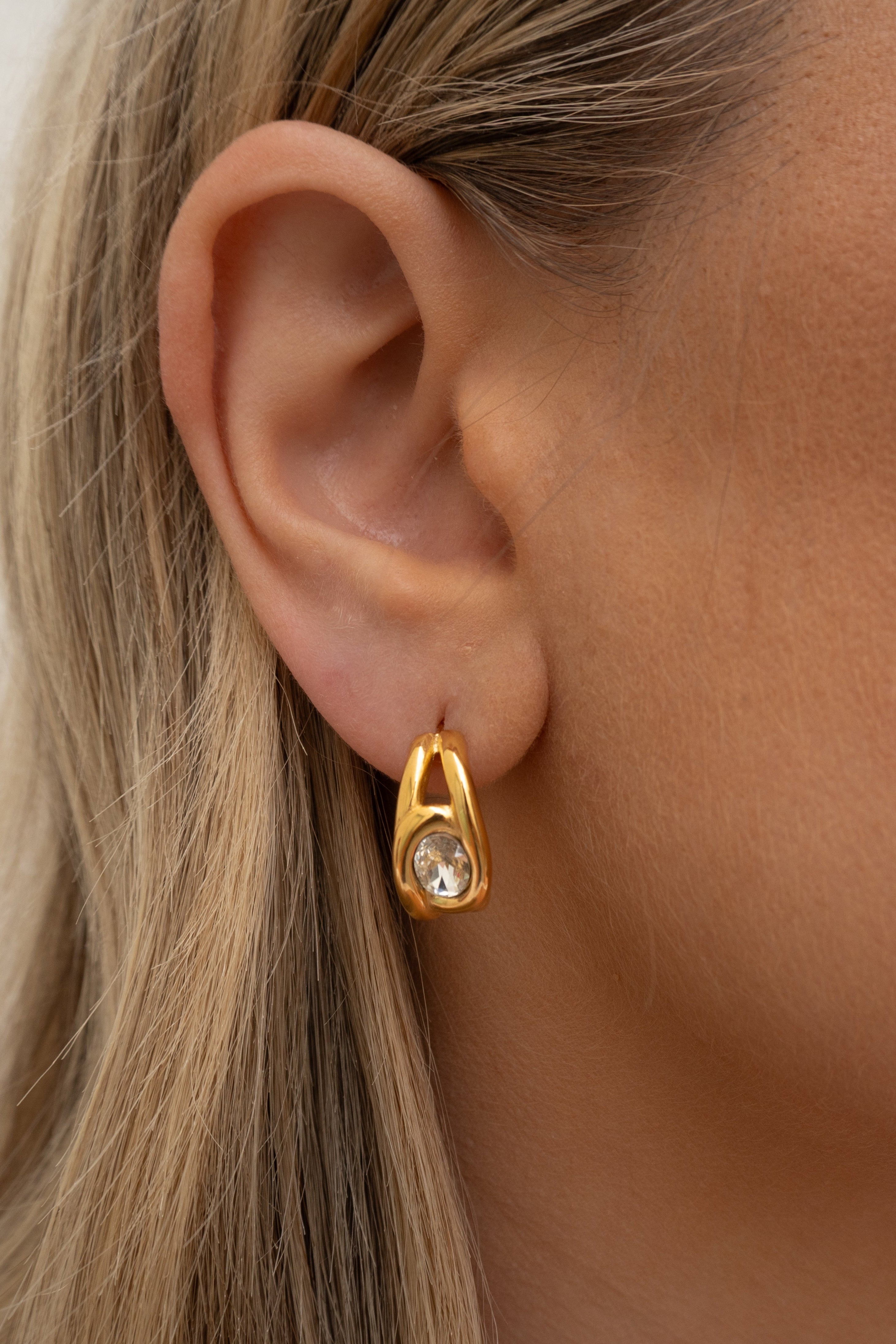 Gold earring with a clear gemstone worn by a person with blonde hair.