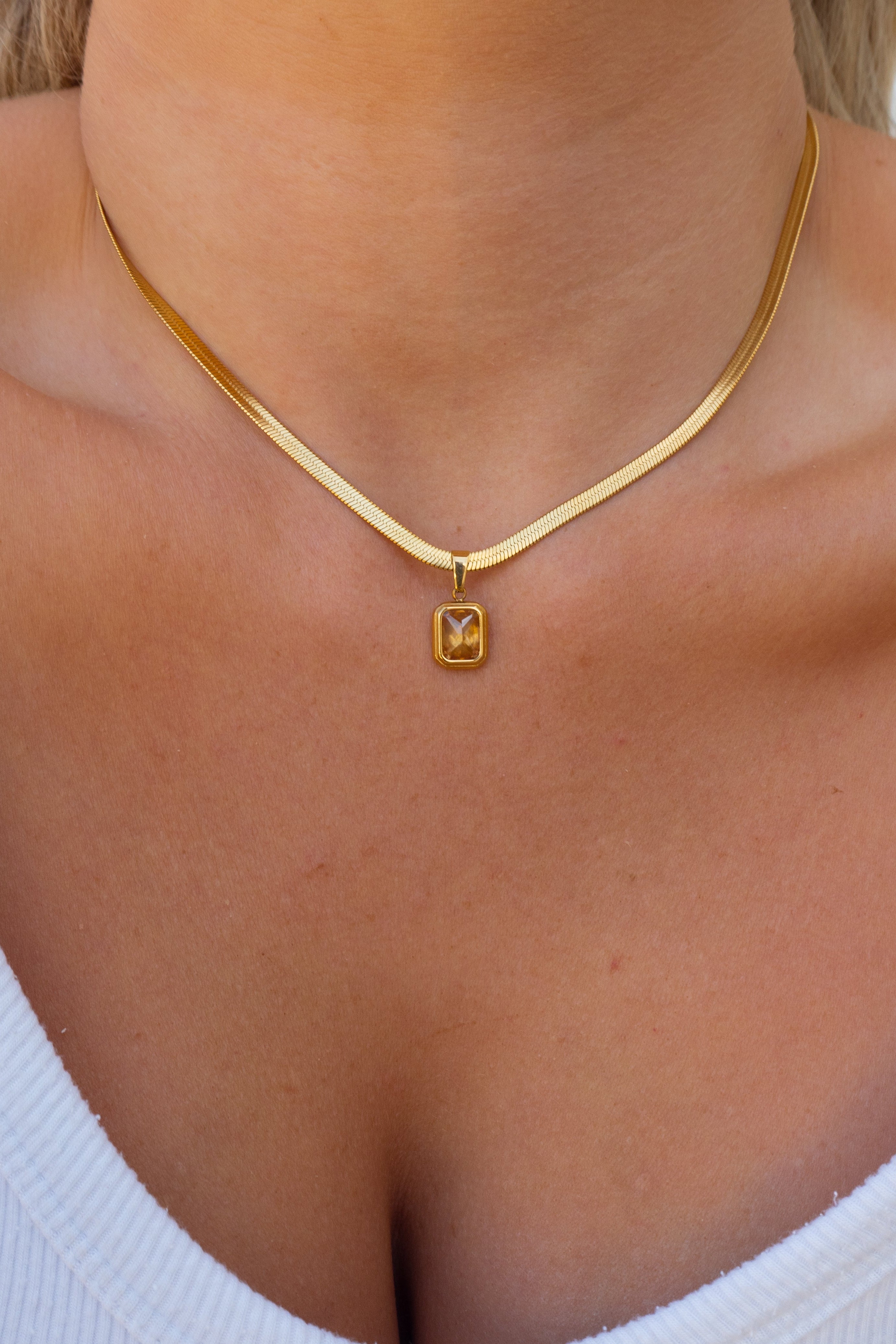 A close-up of a model wearing a gold chain necklace with a small square pendant featuring an engraved letter 'M'.