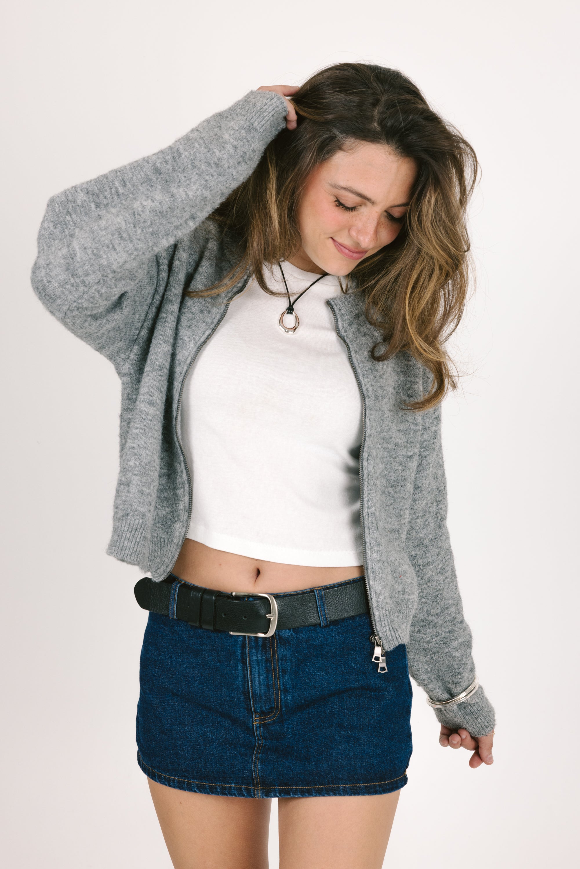 Woman wearing a gray cardigan, white top, and blue denim skirt on a plain background