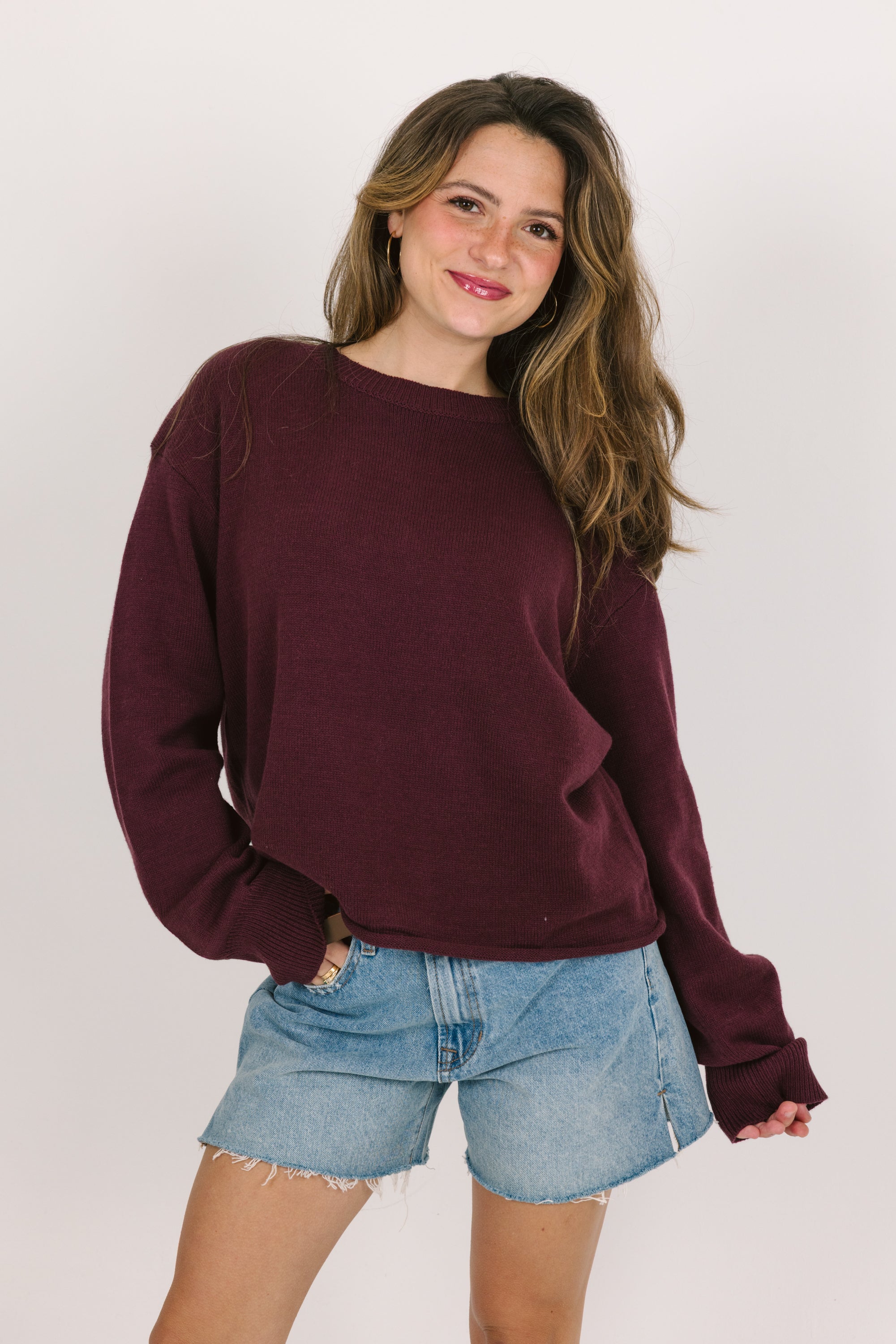 Woman wearing a cranberry sweater and denim shorts on a plain background