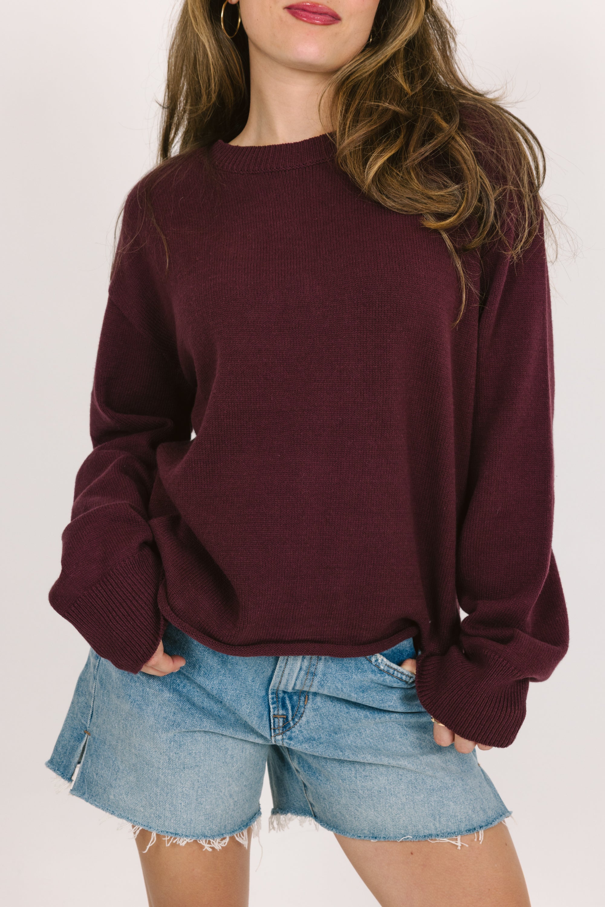 Person wearing a cranberry sweater and denim shorts on a plain background