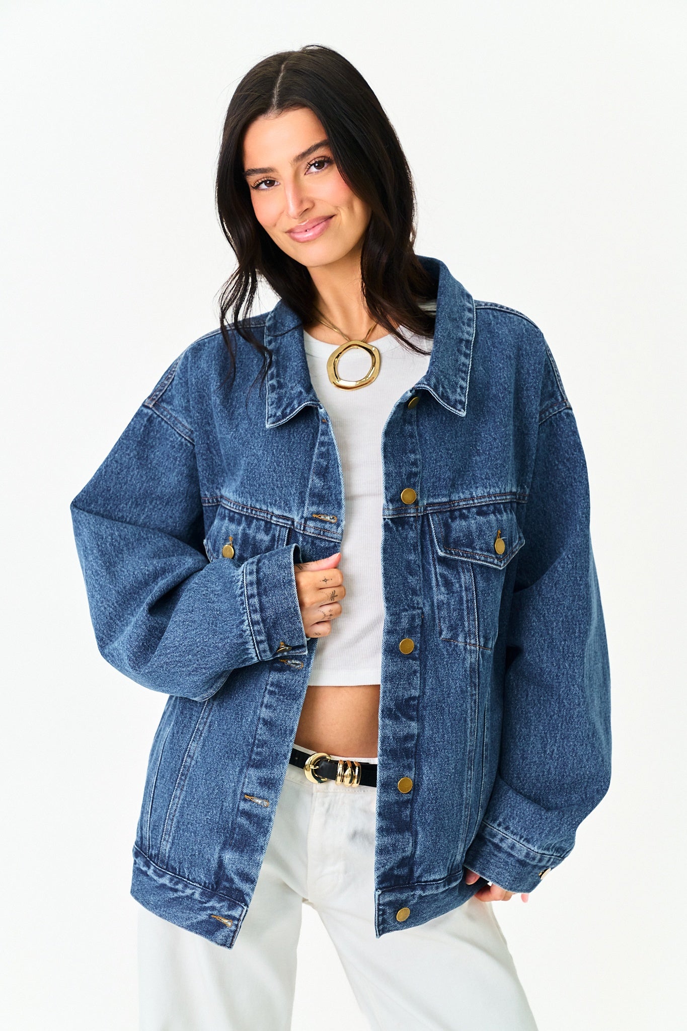 A woman modeling an oversized blue denim jacket over a white crop top and white pants, photographed against a clean white studio background.