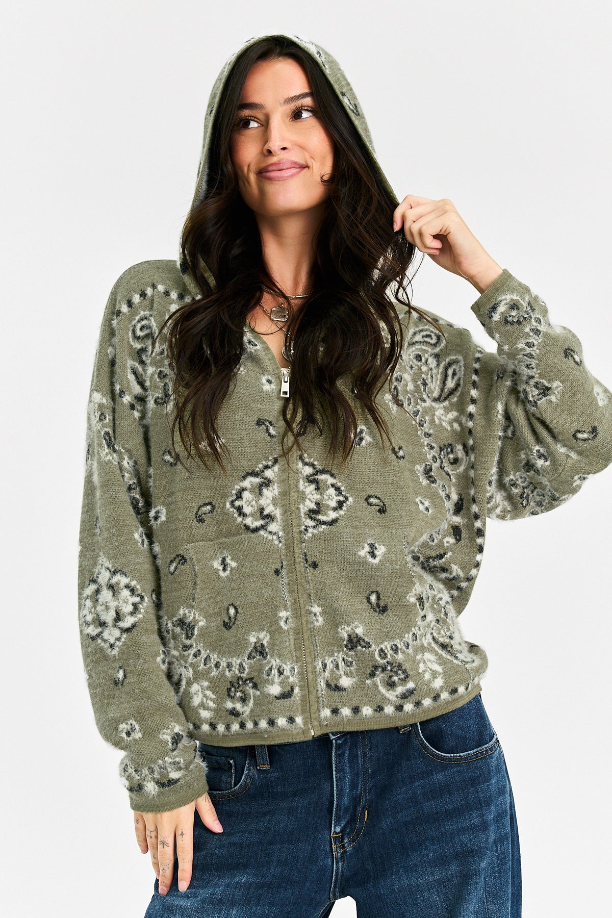 A woman wearing a sage green zip-up hoodie with a gray and black paisley pattern, paired with blue jeans, against a light gray studio background.