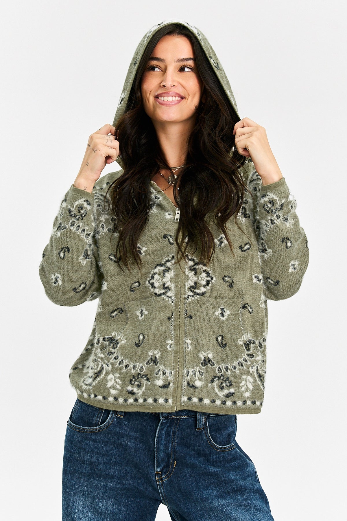 A woman wearing a sage green zip-up hoodie with a gray and white paisley pattern, paired with blue jeans, against a light gray background.