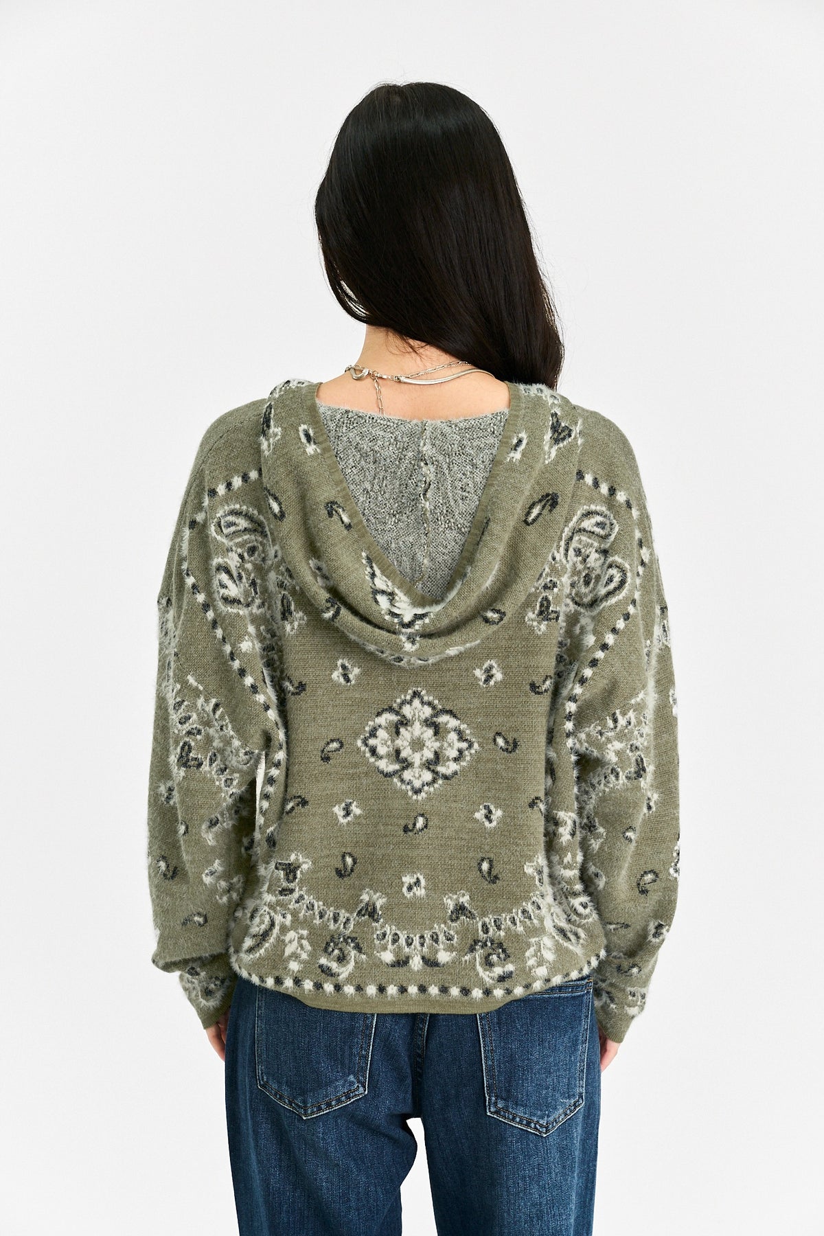 A woman viewed from behind wearing a textured olive green cowl-neck sweater with a gray knit inner hood and paisley-style patterns, paired with blue jeans against a plain white background.