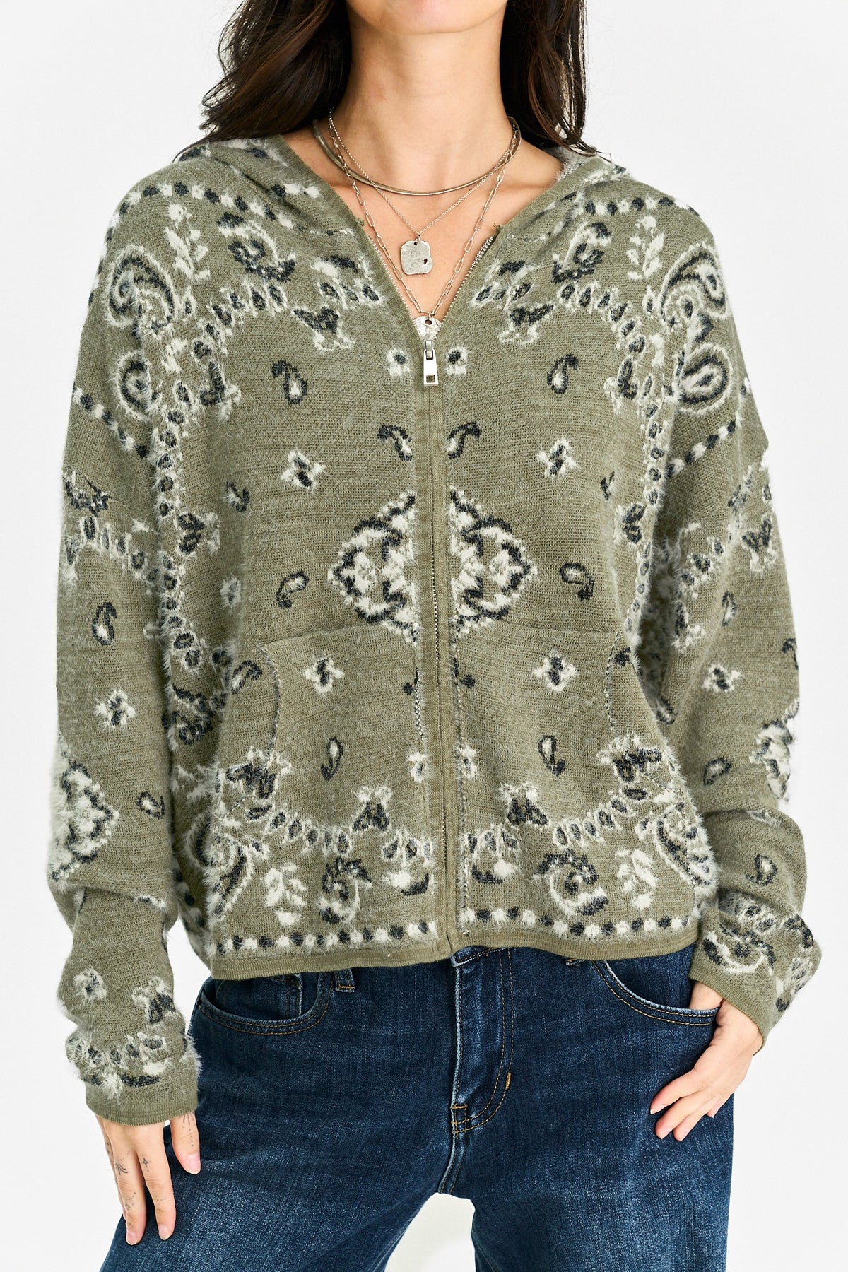 A woman wearing a zip-up hooded sweater with a gray and white paisley pattern on an olive green base, paired with dark blue jeans.