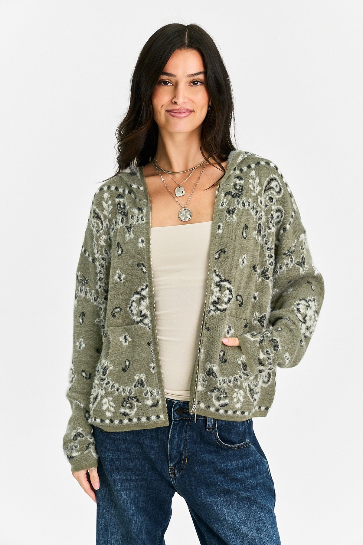 A woman modeling a sage green knit cardigan with a black and white paisley pattern, worn open over a cream tank top and blue jeans against a light gray studio background.